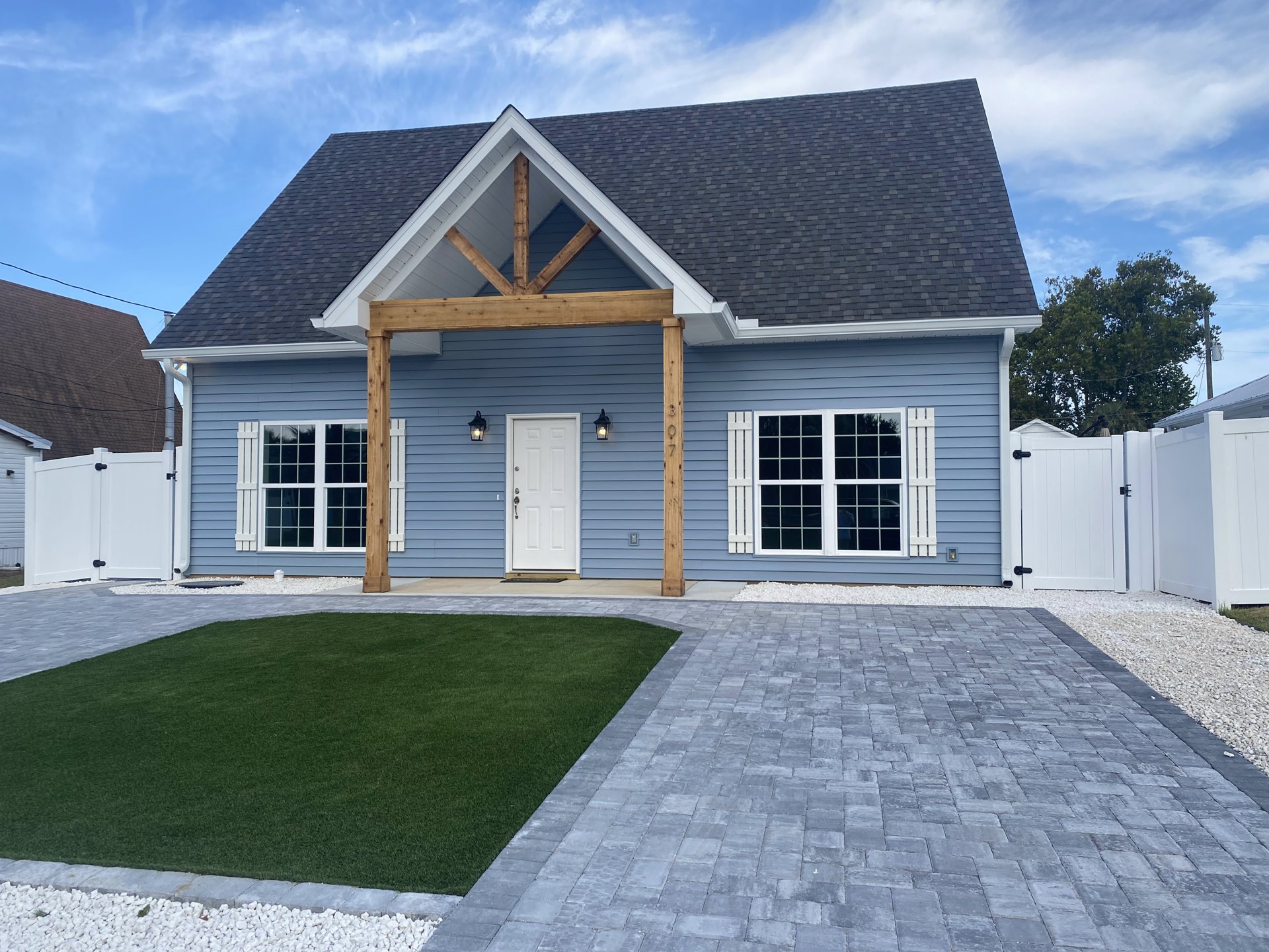 Two-story cottage with white siding, multiple square windows, white front door with silver handles, wooden arch over stone walkway, grey brick patio, green lawn, and white picket