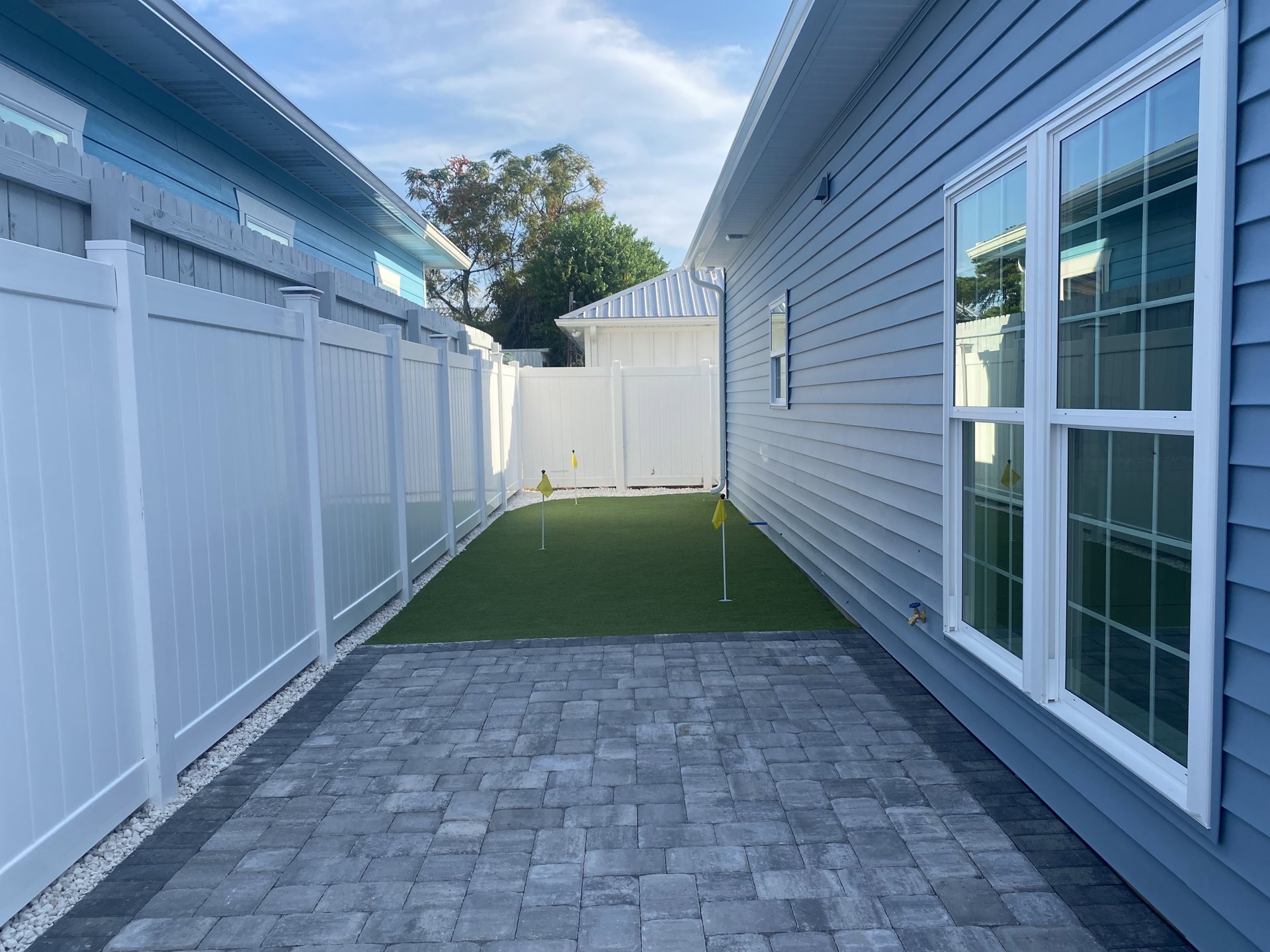 Stone patio bordered by green lawn, white picket fence, glass-paned window, and tree in the background with a small flag on the grass
