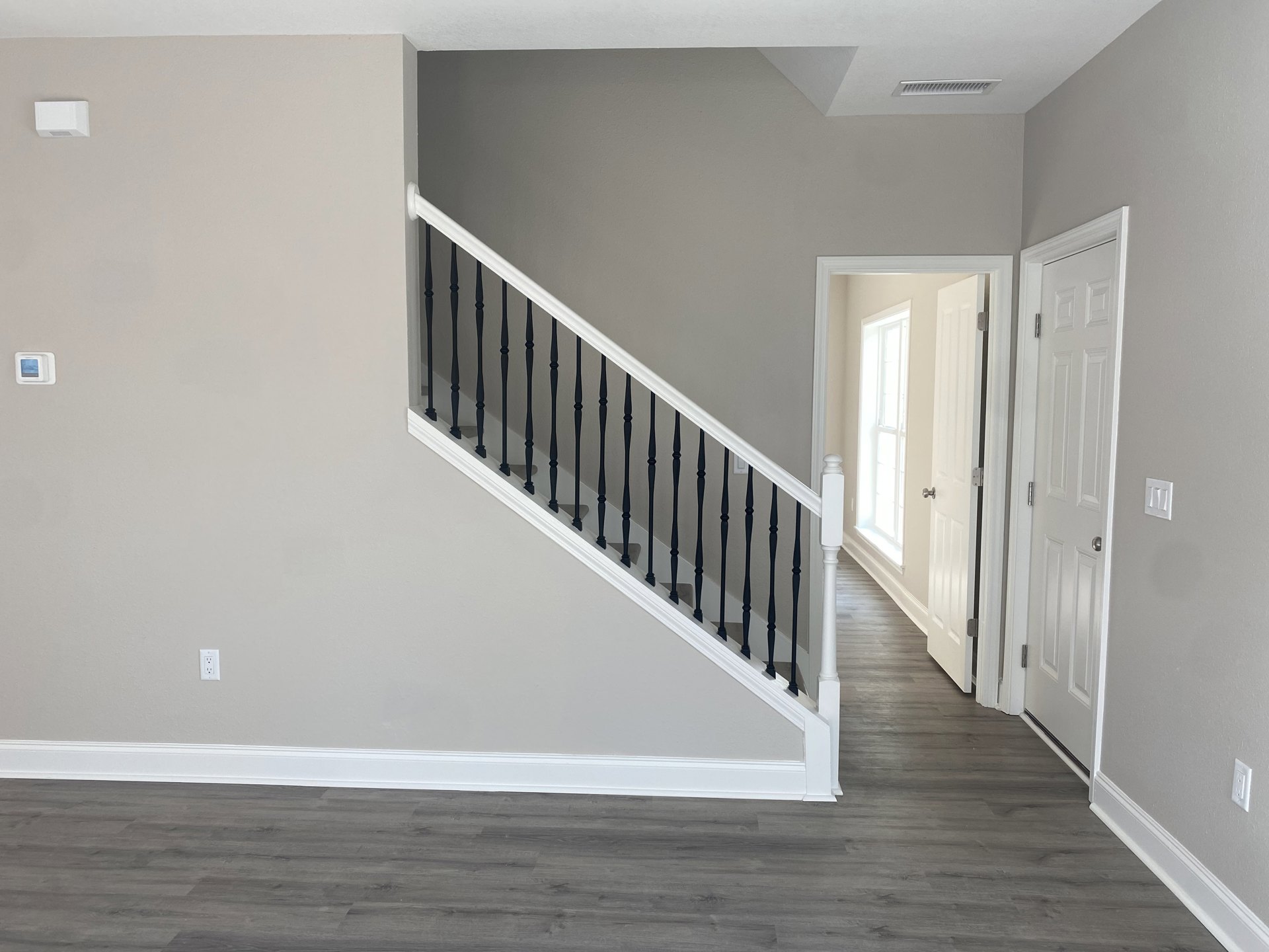 Wood staircase with black metal railings, white walls, wood flooring with white baseboards, white door with silver handle, window letting in natural light, white vent box on wall