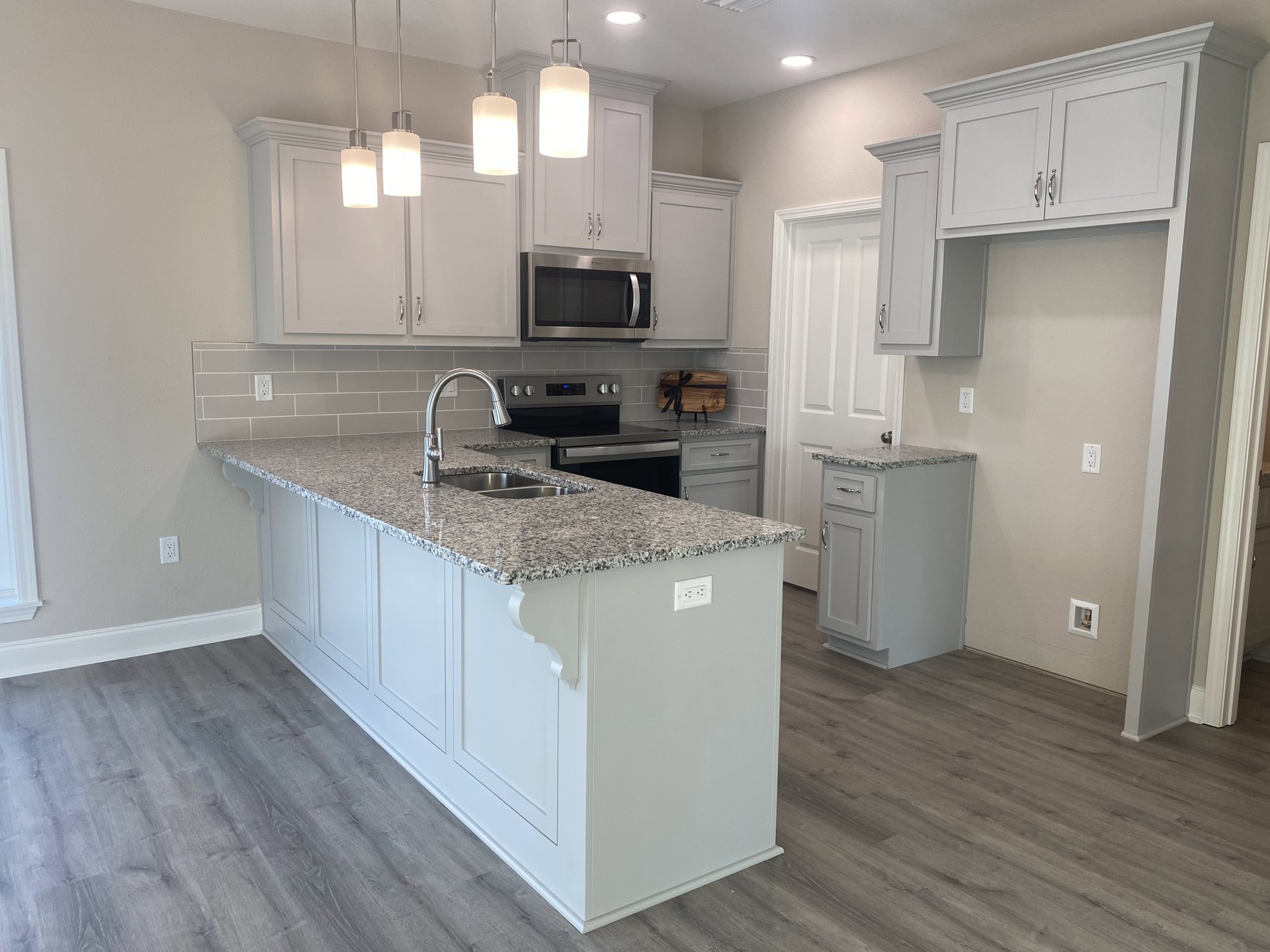 Marble countertop kitchen with wood flooring, white cabinetry, stainless steel microwave, island featuring sink and stove, pendant light, and wooden cutting board with black ribbon
