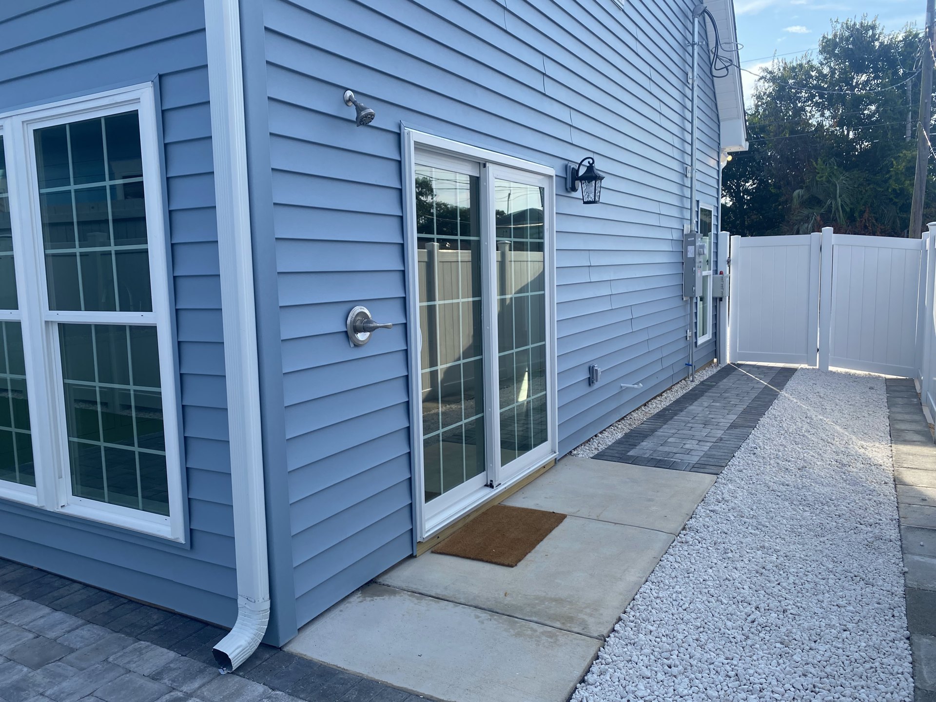 Blue house exterior with white gate, sliding glass door, multiple windows, brown doormat on concrete, and water spout near entry.