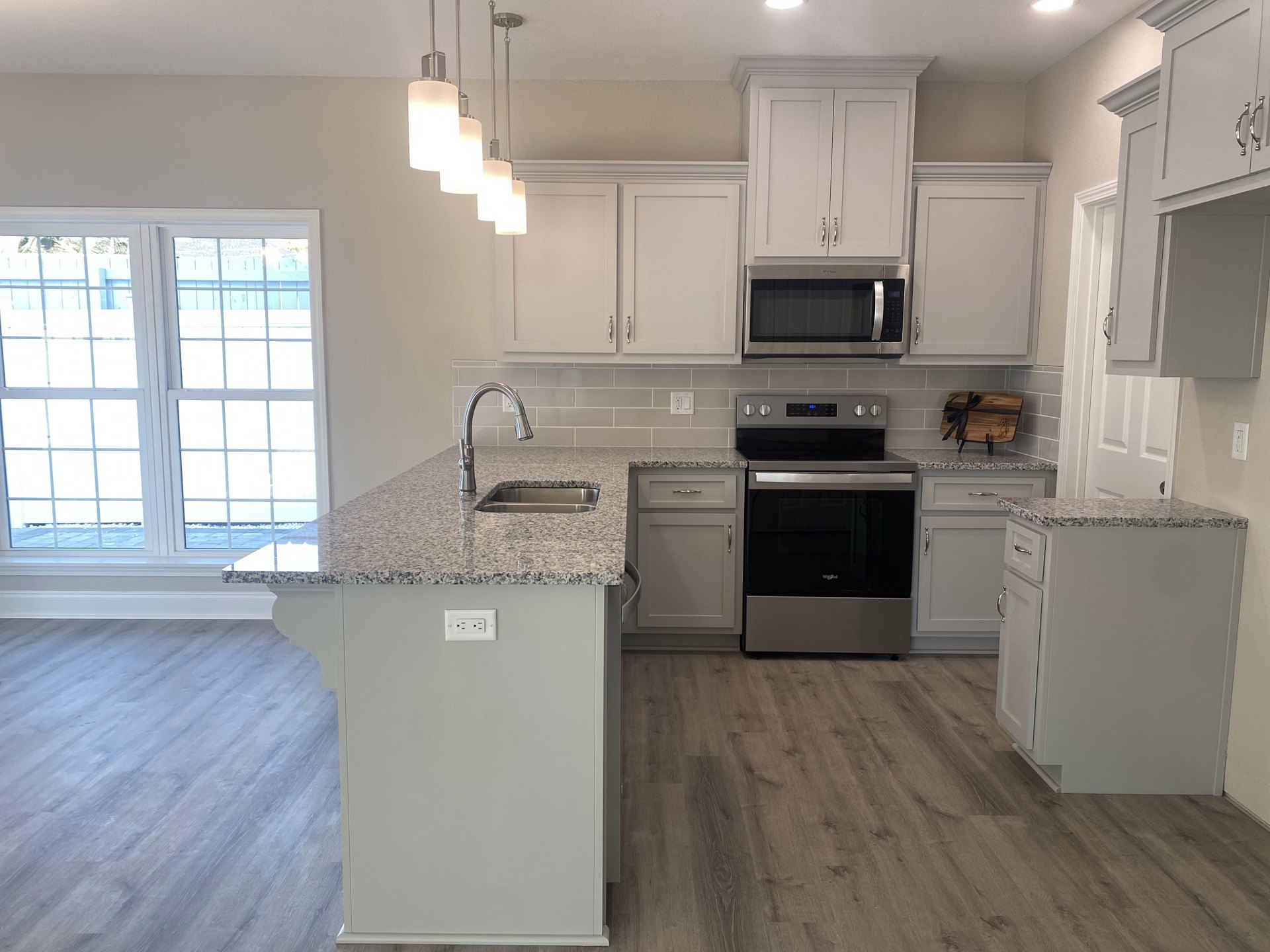 White kitchen with granite countertops, black and silver oven, built-in microwave, multi-pane window, wooden cutting board with black ribbon, white island topped with marble.