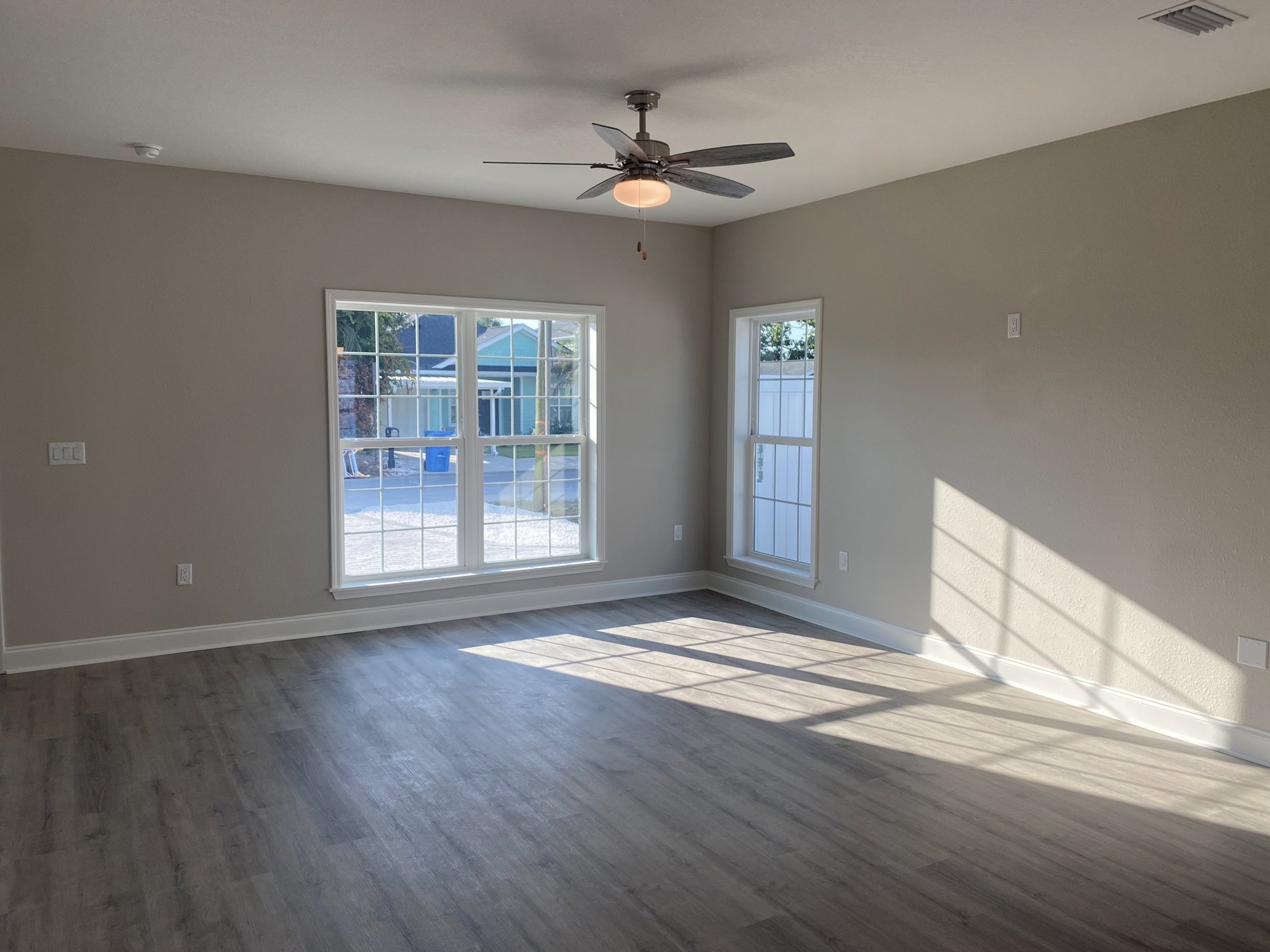 Living room with wood flooring, white plaster walls, ceiling fan with light, and multiple windows showing views of a snowy street and blue rooftops.