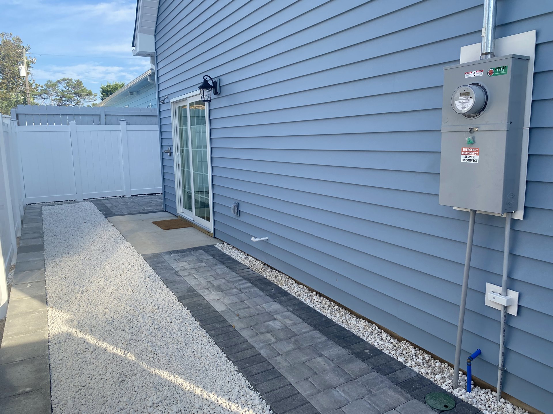 Two-story house with blue siding, white trim, attached garage, white picket fence, sliding glass door, grey electrical meter with warning sign, and outdoor lamp.