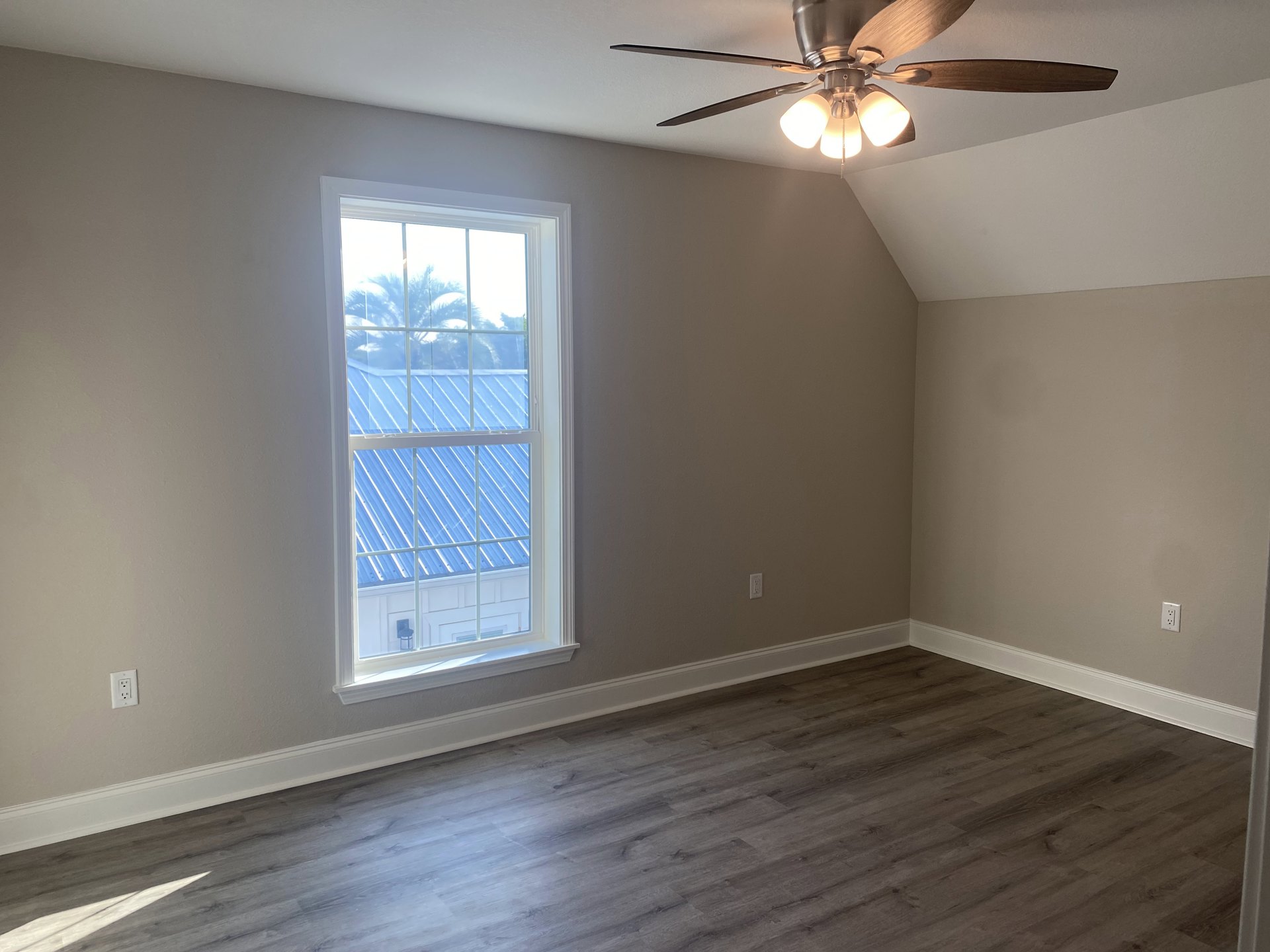 Ceiling fan with light fixture mounted on white plaster ceiling, large window framed by palm trees outside, wood laminate flooring with sunlight casting shadows across the room.
