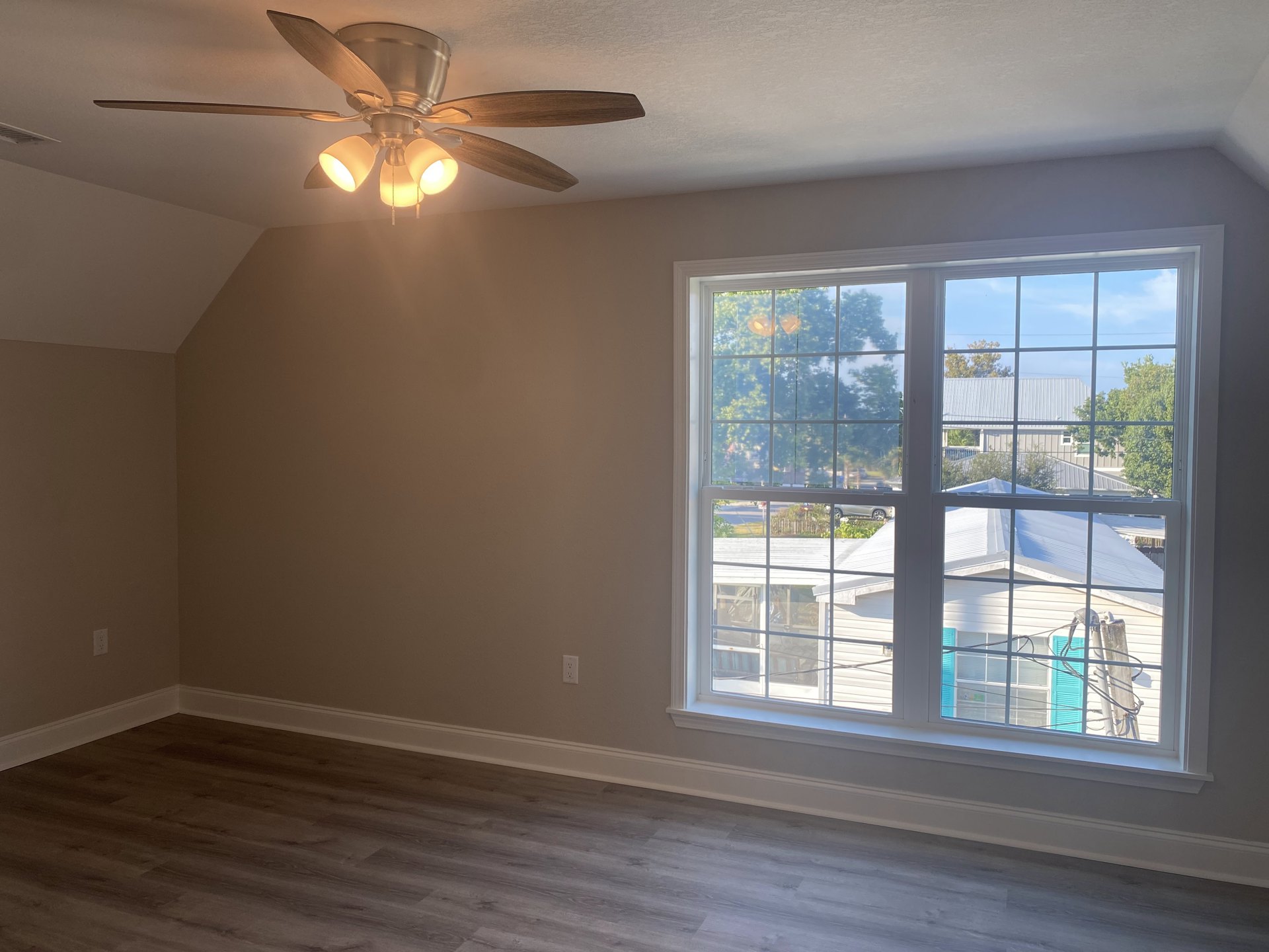 Ceiling fan with light fixture mounted on white plaster ceiling, wood floor beneath, blue shuttered window with white trim offering view of neighboring house and trees.