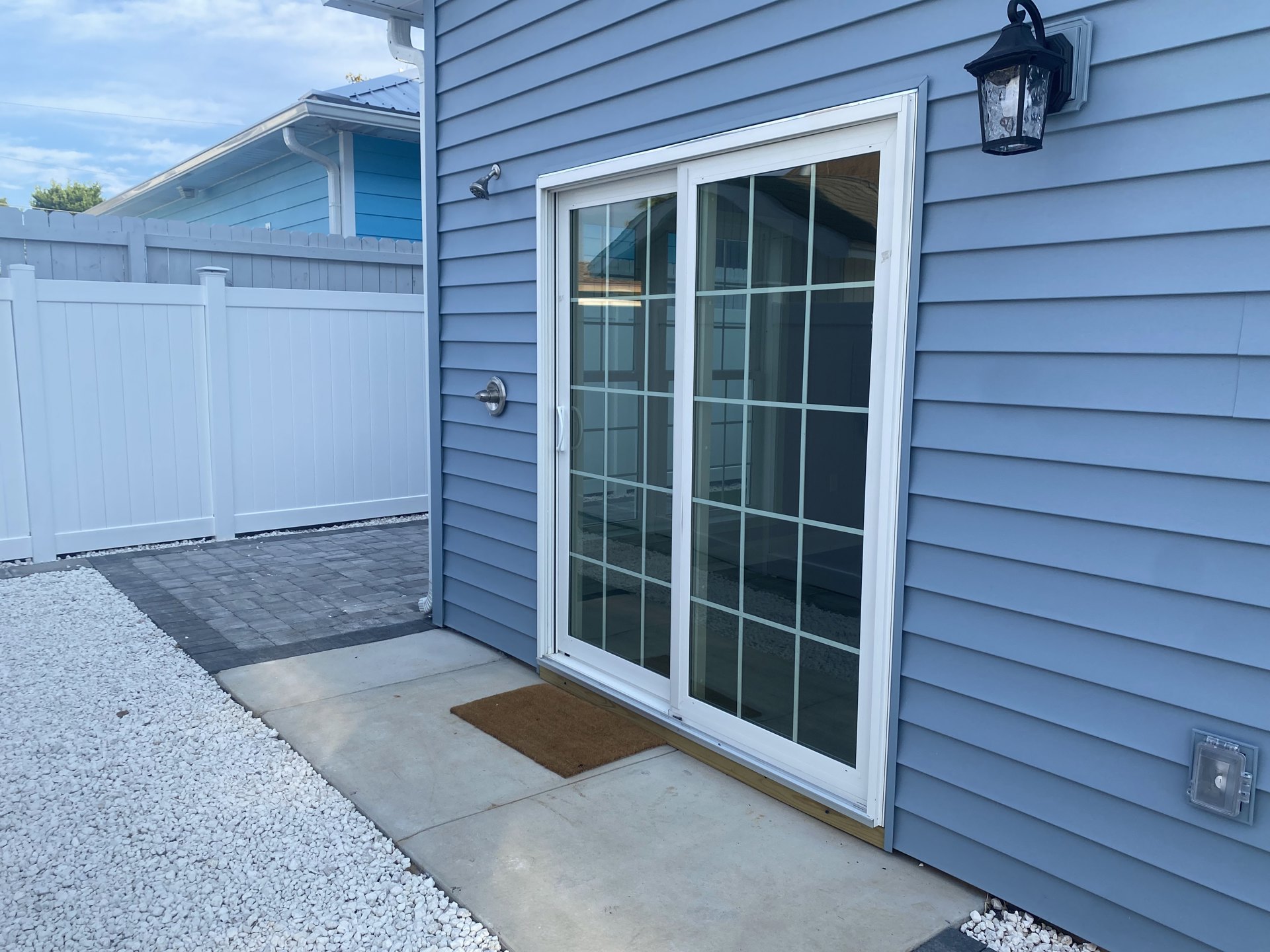 Brown front door with glass panes, black wall-mounted light fixture, plastic key box, brown doormat on white porch surface, white gravel beside red brick walkway, white siding