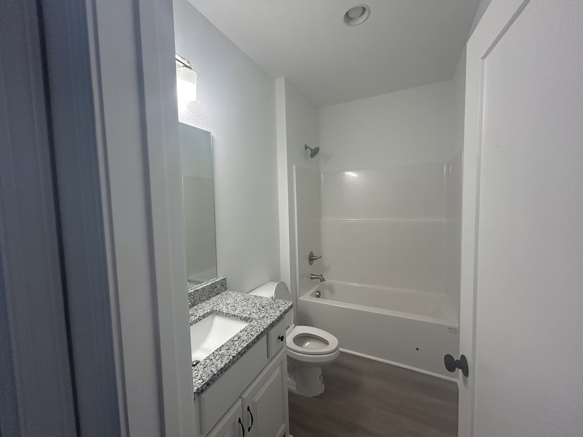 Bathroom with white toilet and bathtub, black faucet fixtures, white sink with black speckled countertop, wood flooring, and white baseboards