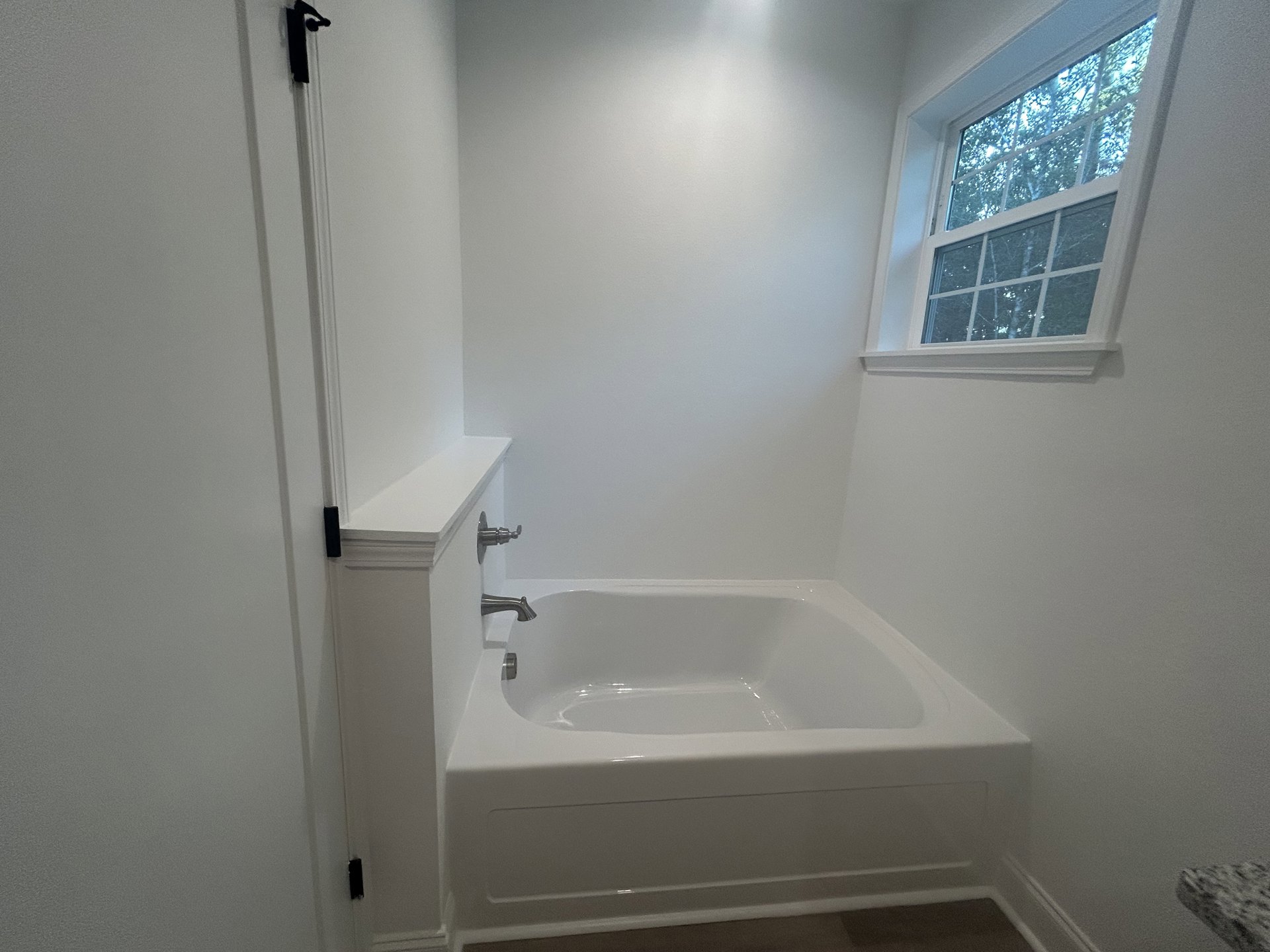 Freestanding white bathtub beneath a window with leafy trees outside, chrome faucet, light reflecting on tub surface, white tile walls, built-in shelf