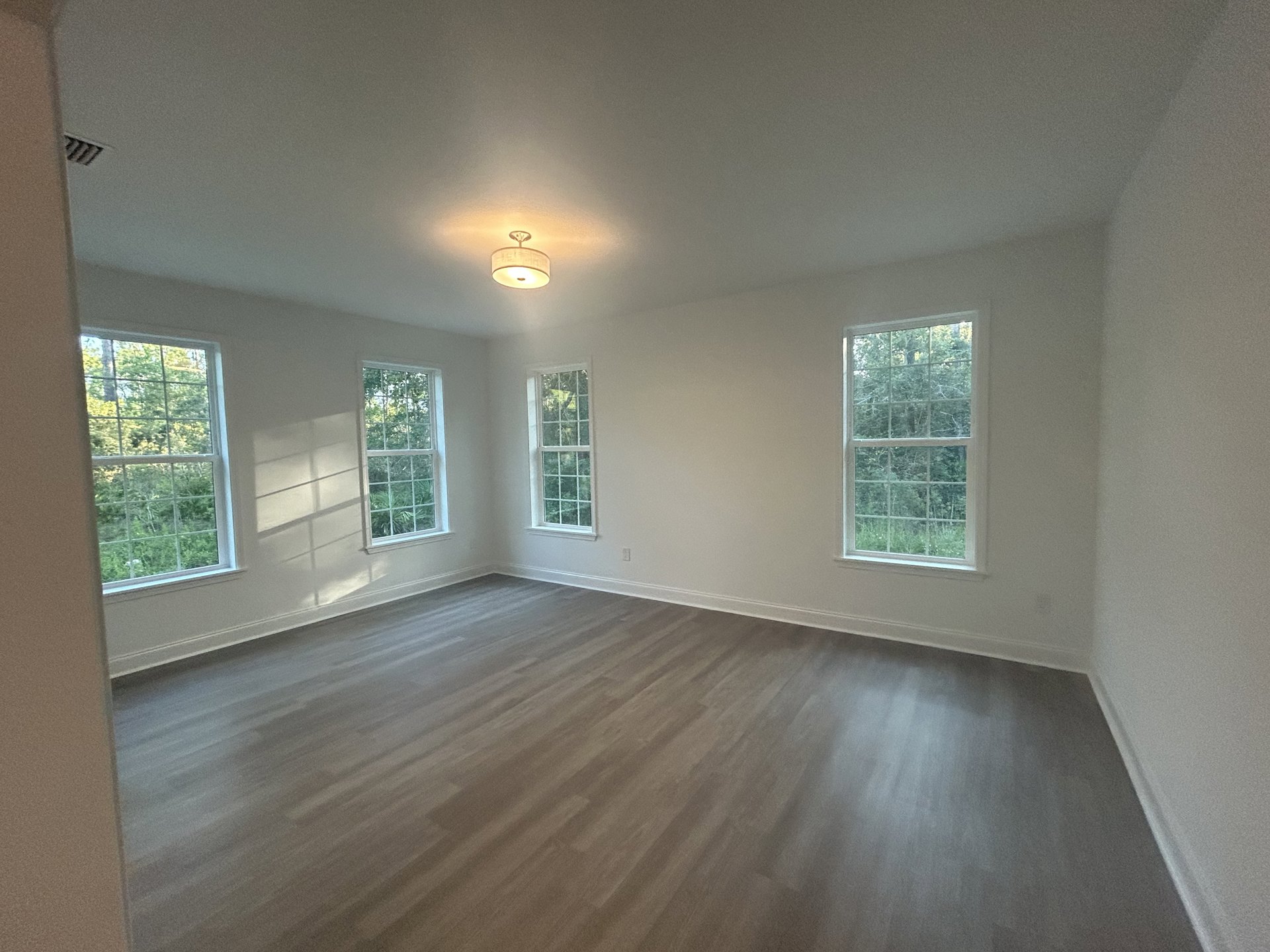 Sunlit room featuring multiple white-framed windows overlooking trees, hardwood flooring, pale plaster walls, and a ceiling light fixture with a white shade