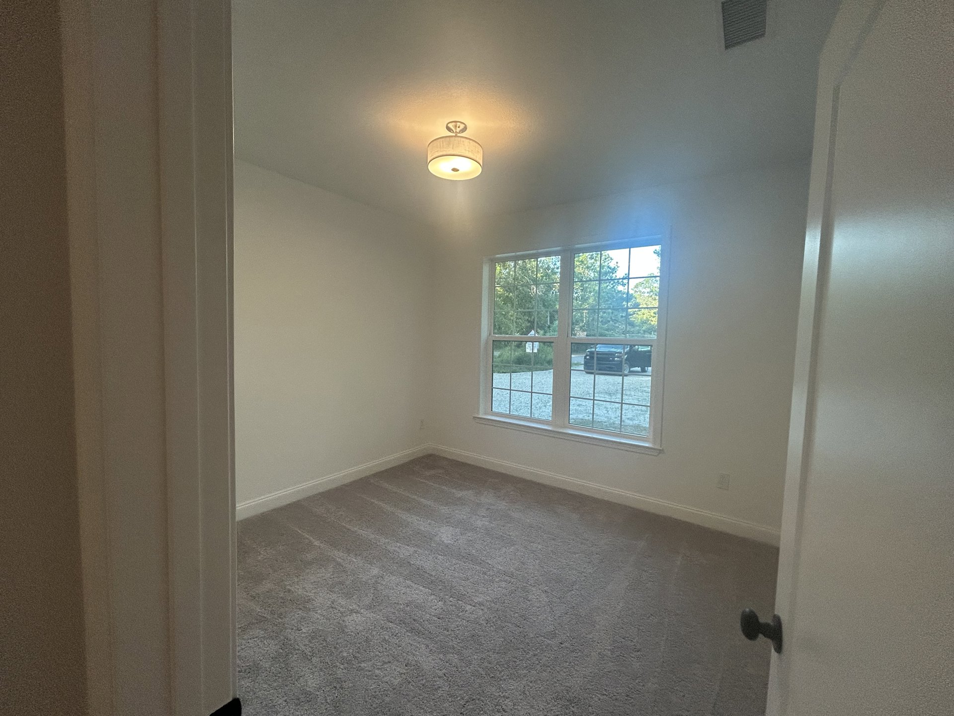 Neutral-toned room with carpet flooring, white plaster walls, large window revealing parked car outside, and ceiling-mounted light fixture with white shade