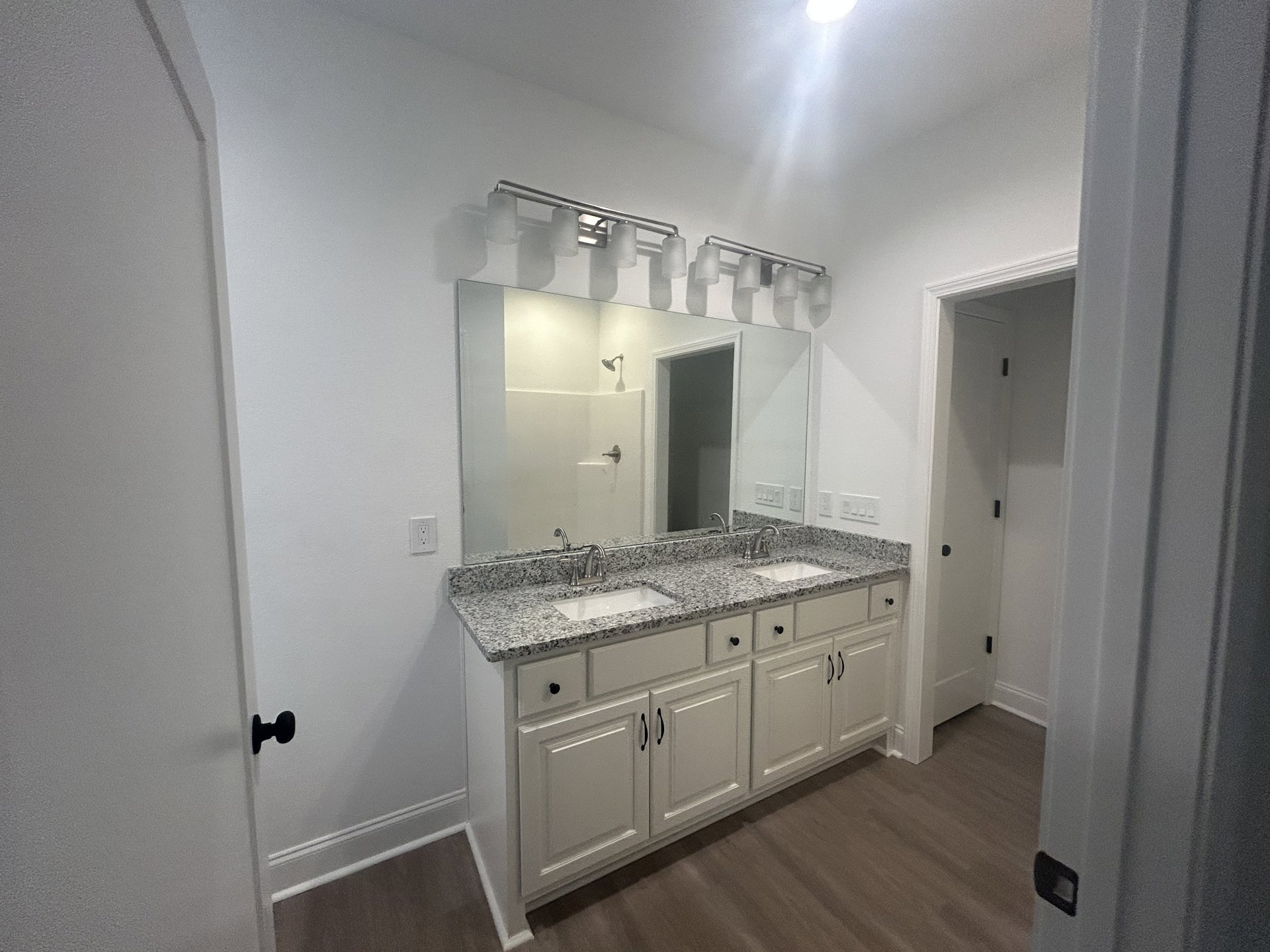 White bathroom with expansive mirror above dual sinks, white cabinetry, black door knob on white door, and four-shade light fixture mounted above mirror