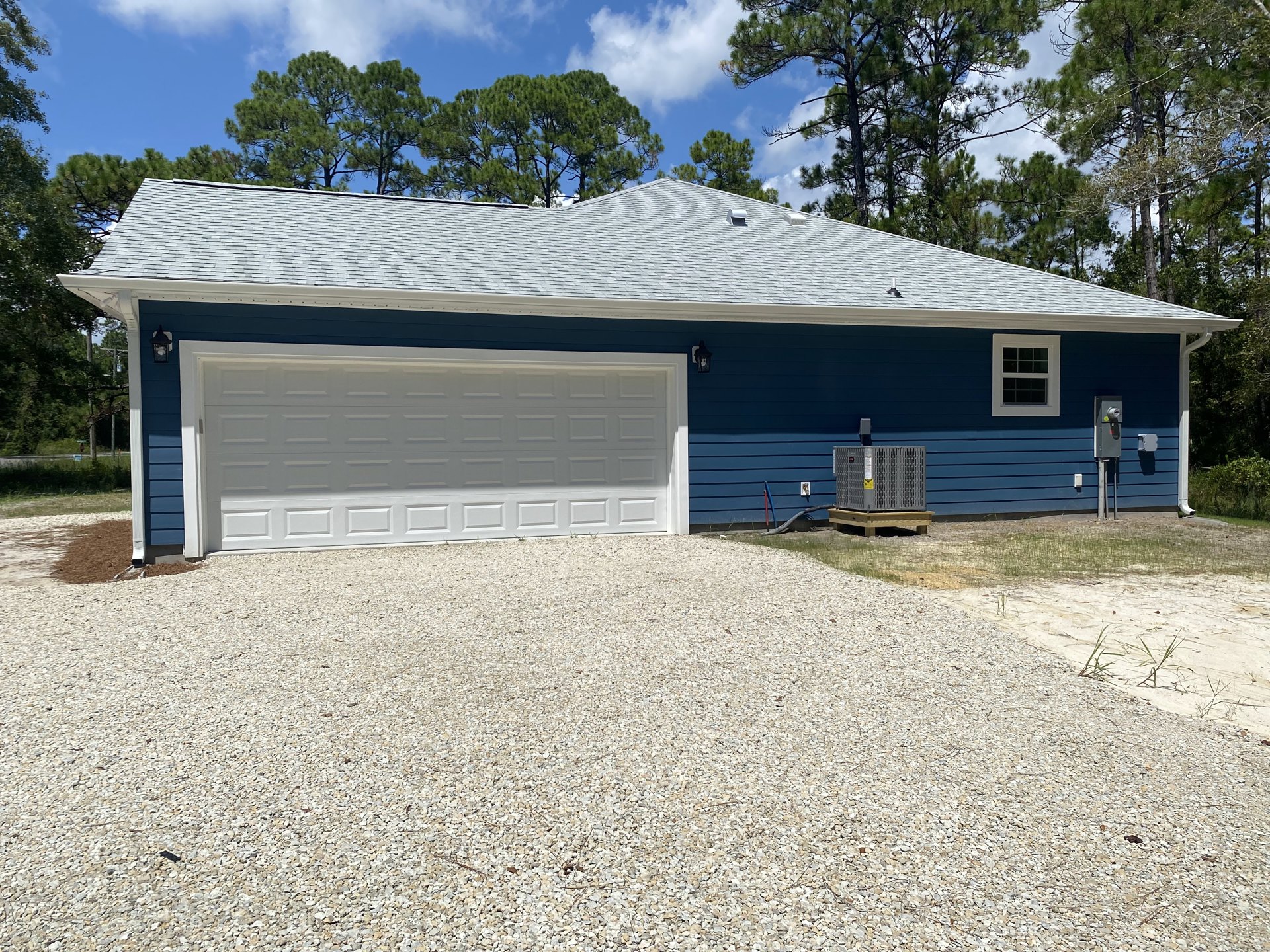 Blue siding house with white-trimmed window, black roof, attached garage featuring white door and hose, gravel driveway, trees in background.