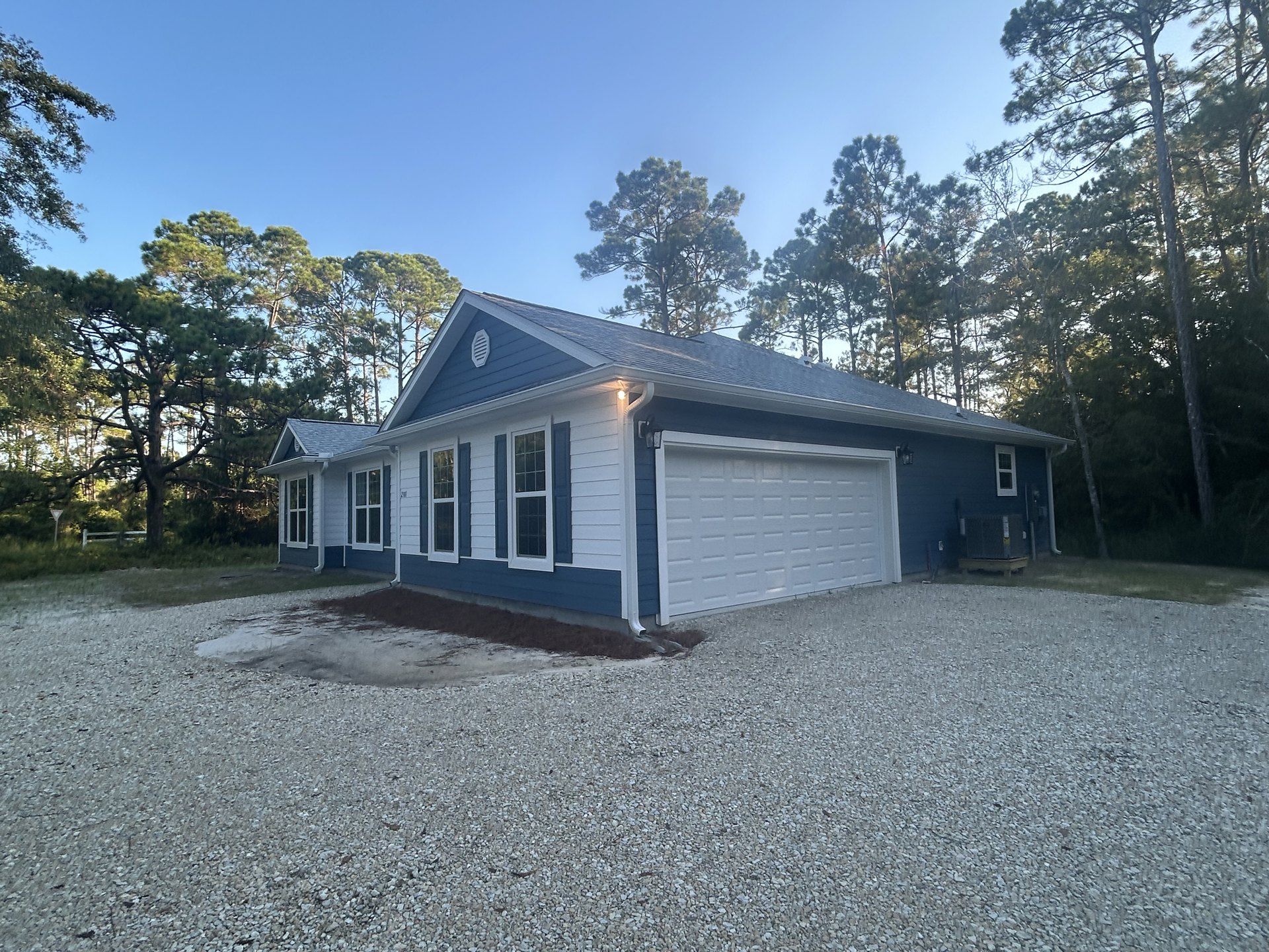 Two-story house with white garage door, black roof, gravel driveway, white-framed windows, surrounded by tall trees in the background