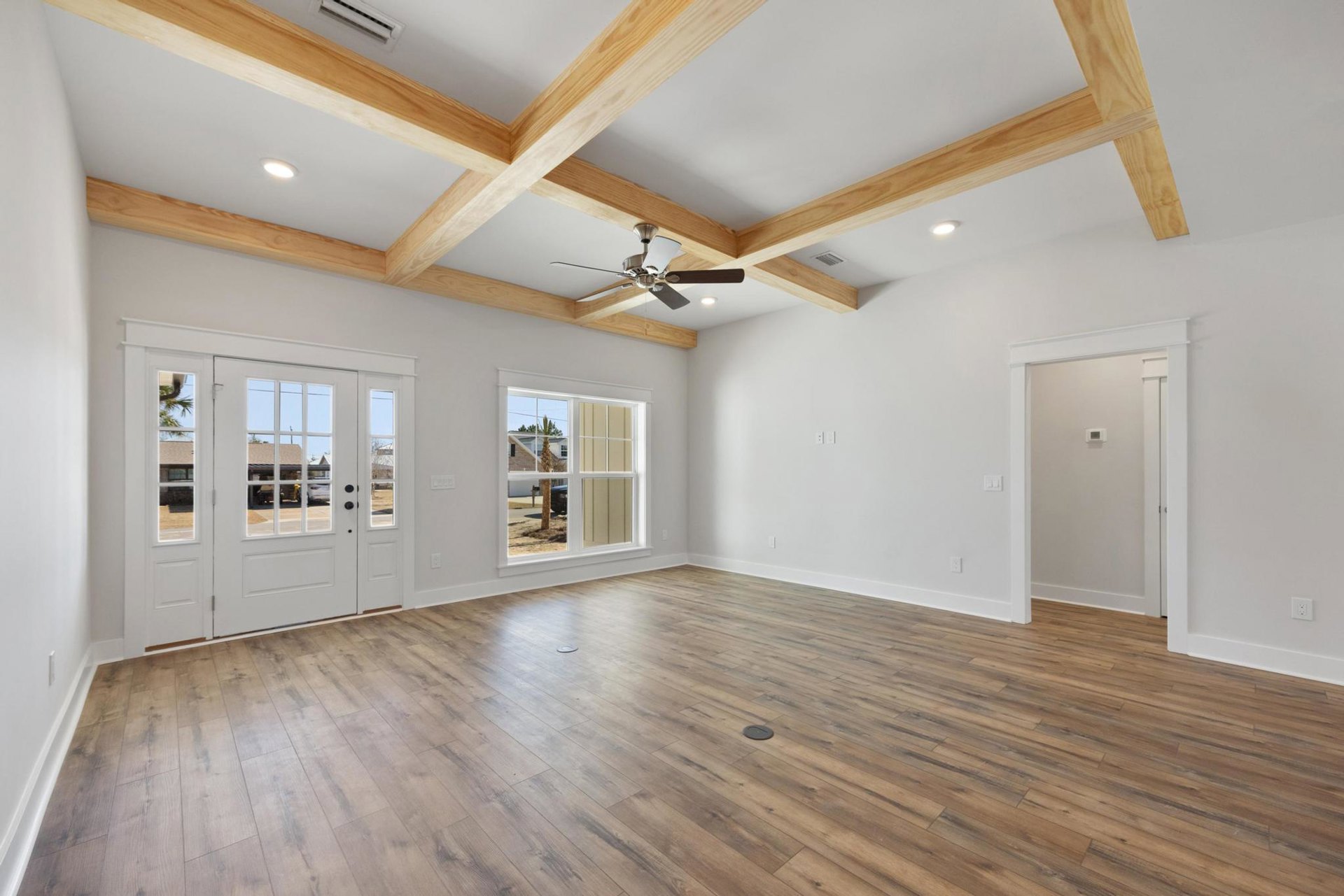 Spacious room with hardwood floors, white double doors with glass panes, ceiling fan mounted on wooden beam, neutral walls, and thermostat near entry.