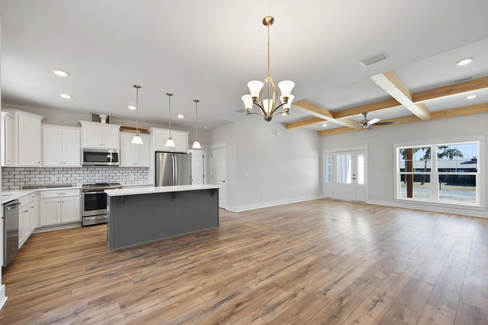 Open kitchen and living room with wood flooring, white walls, grey accent wall, white countertops, stainless steel refrigerator, oven, and stove, large window overlooking