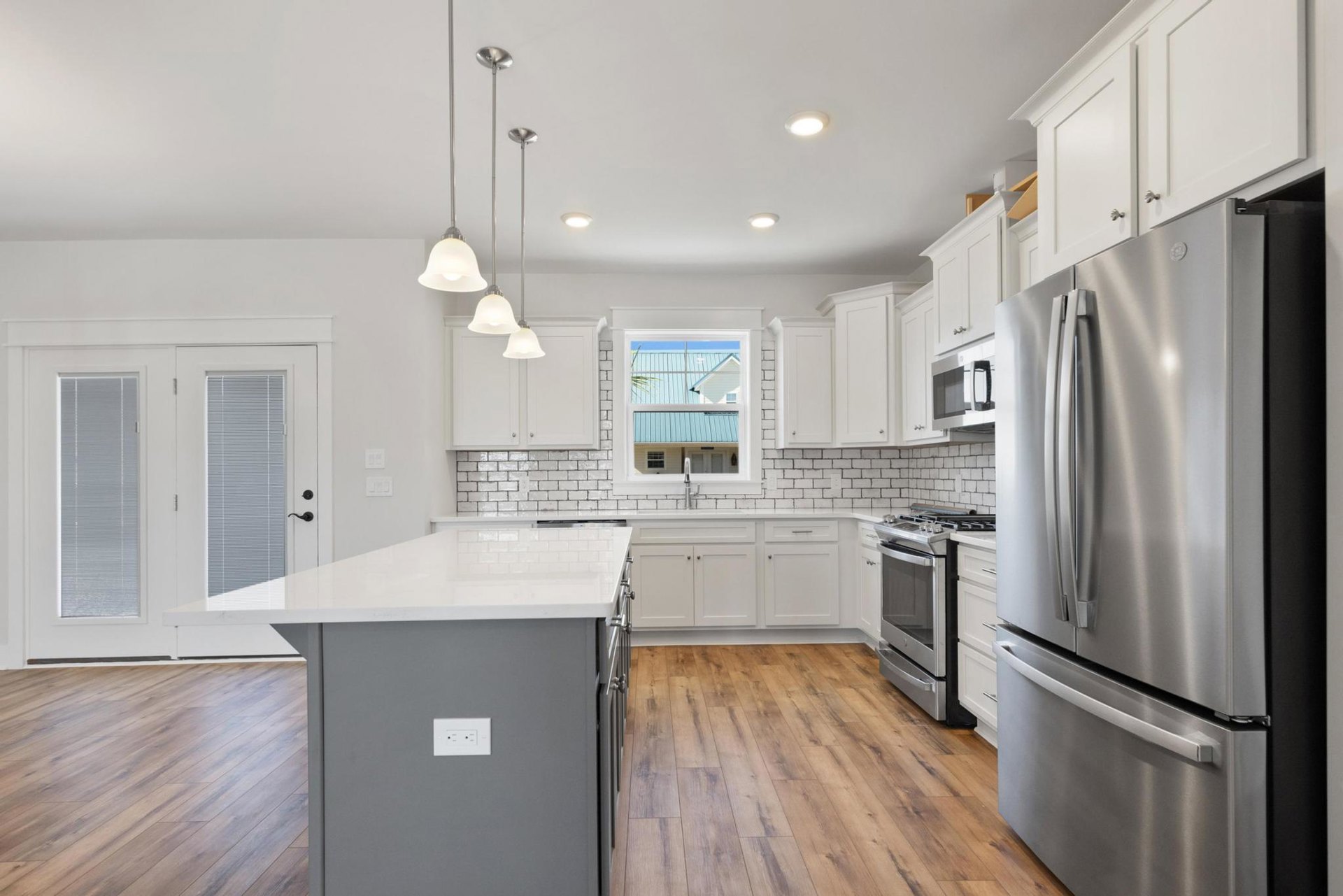 White kitchen cabinets and a grey island with quartz countertops, stainless steel refrigerator, white electrical outlets, large window showing a neighboring house and palm tree