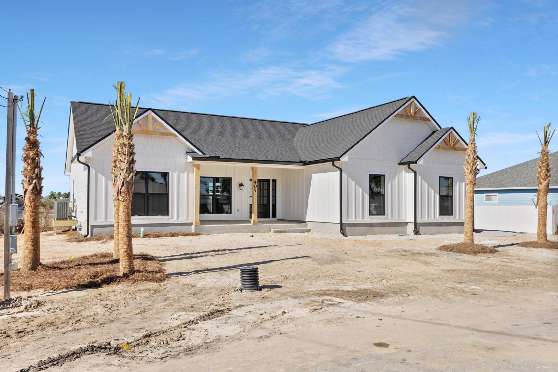 Modern stucco house with black-framed windows, palm tree planted in front yard, exposed black irrigation pipe in soil, cloudy sky overhead