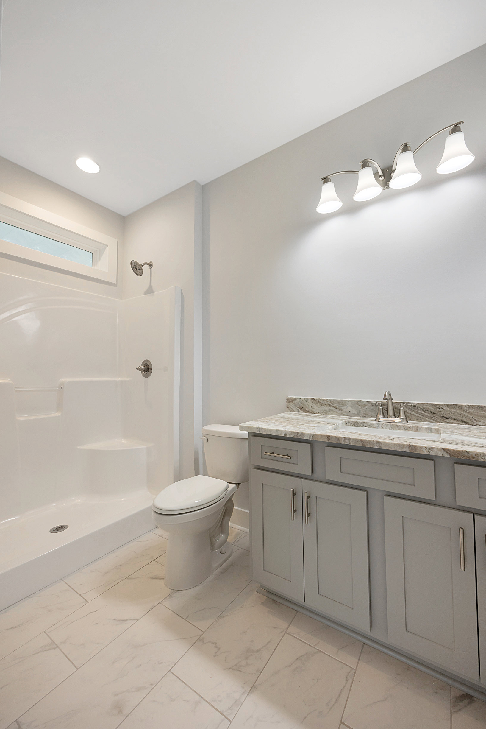 Marble-topped vanity with undermount sink, three-light fixture above mirror, white toilet with closed lid, light-colored tile flooring, neutral painted walls