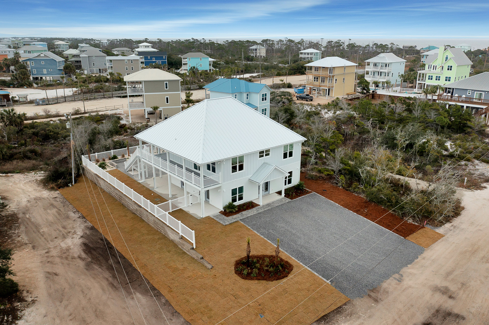 White house with white roof and fence, gravel driveway, palm trees in landscaped circular area, building under construction with ladders, blue sky with thin clouds