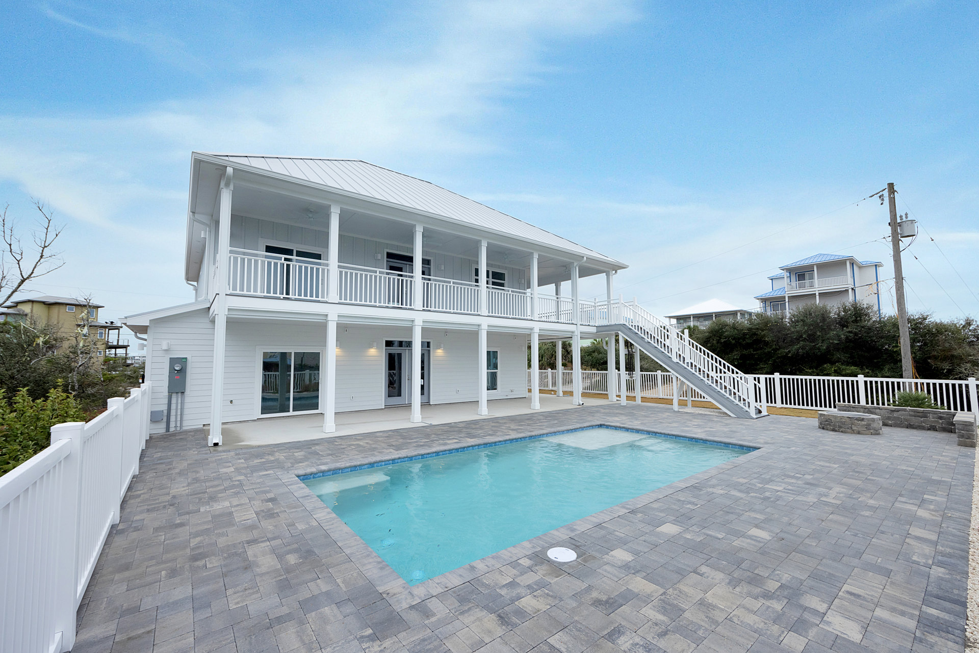 Rectangular swimming pool bordered by stone walkway, white house with balcony and white railing in background, white fence and brick path alongside lush landscaping.