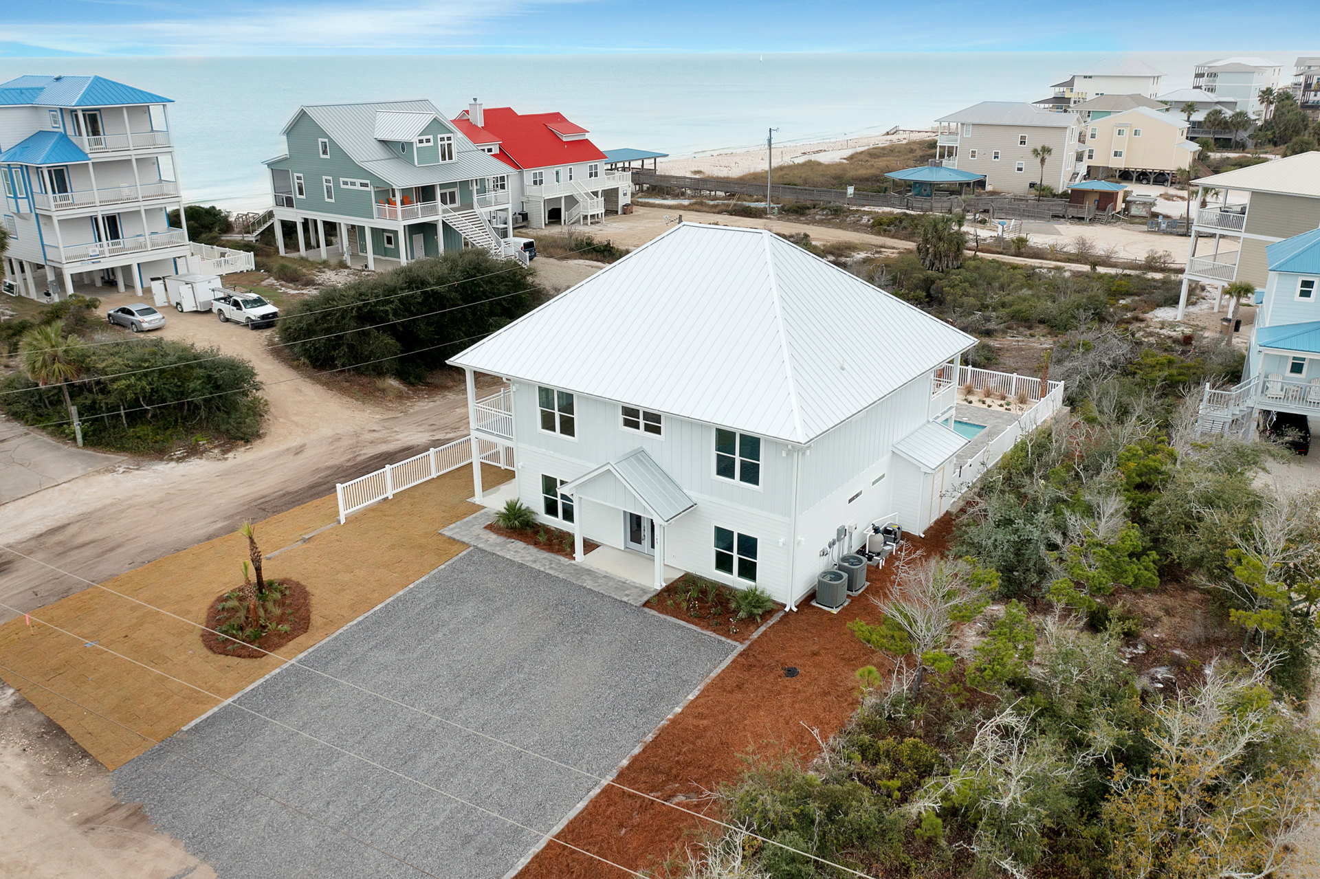 White house with blue roof, palm trees, paved driveway, sandy beach and ocean in the background, partly cloudy sky