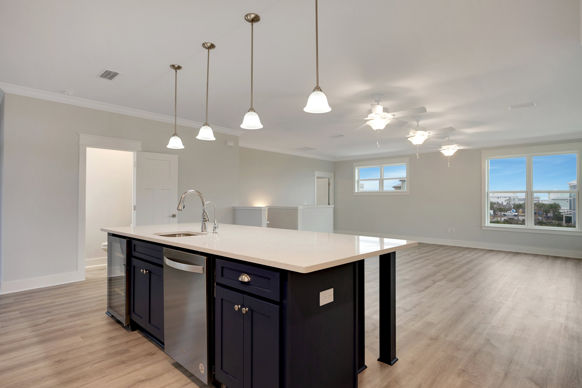 Quartz kitchen island with pendant lights, white cabinetry, stainless steel sink and dishwasher, white electrical outlet, window overlooking street and trees, white ceiling fan