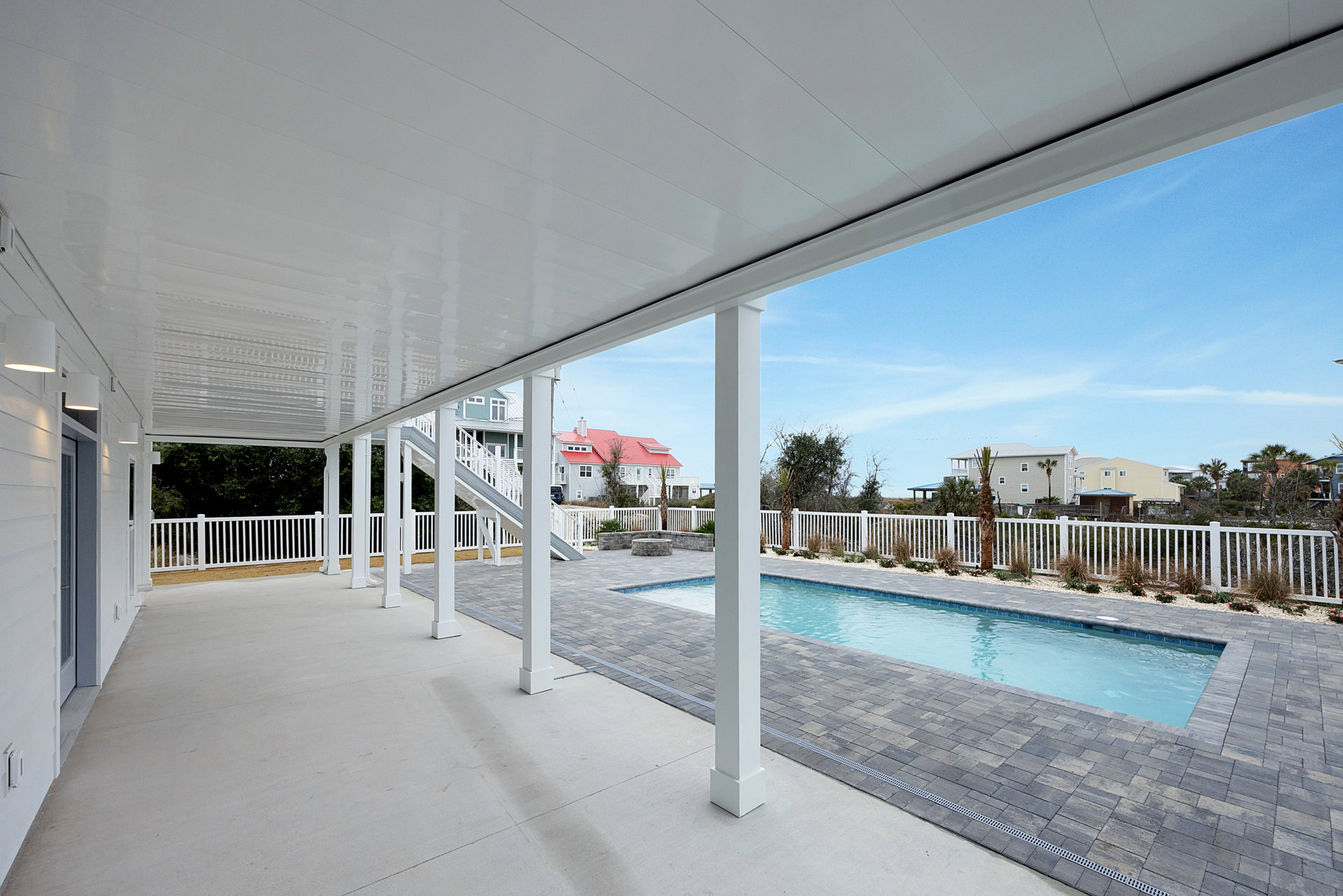 Rectangular swimming pool bordered by composite deck, white fence, and covered patio with white pillars; house features red roof and white exterior.