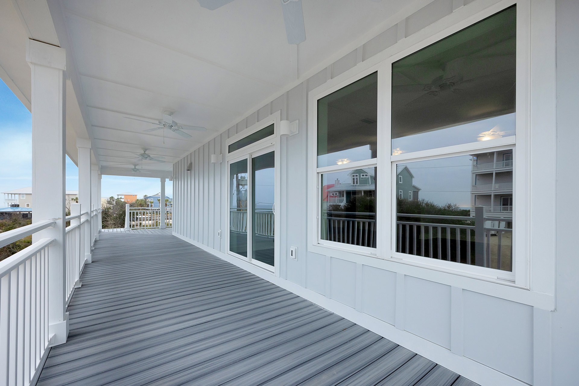 White exterior house with large windows, white vertical railing on porch, grey wood deck flooring, white trim, and blue sky in background