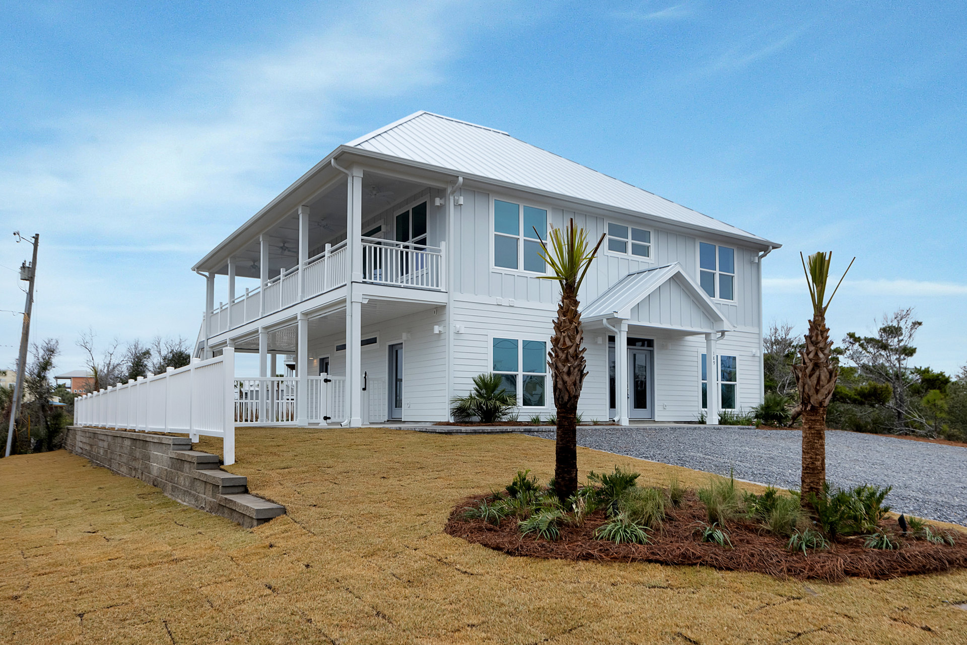 White house with white picket fence, palm trees in front yard, Southfork Ranch visible in background, blue sky with scattered clouds.