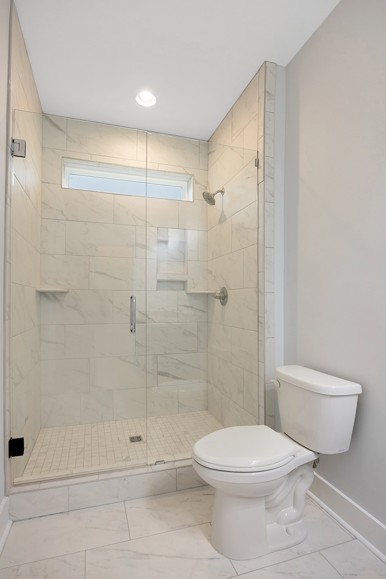Bathroom featuring a frameless glass shower enclosure, white toilet with matching tank, marble wall accents, recessed ceiling light, and a window providing natural light.