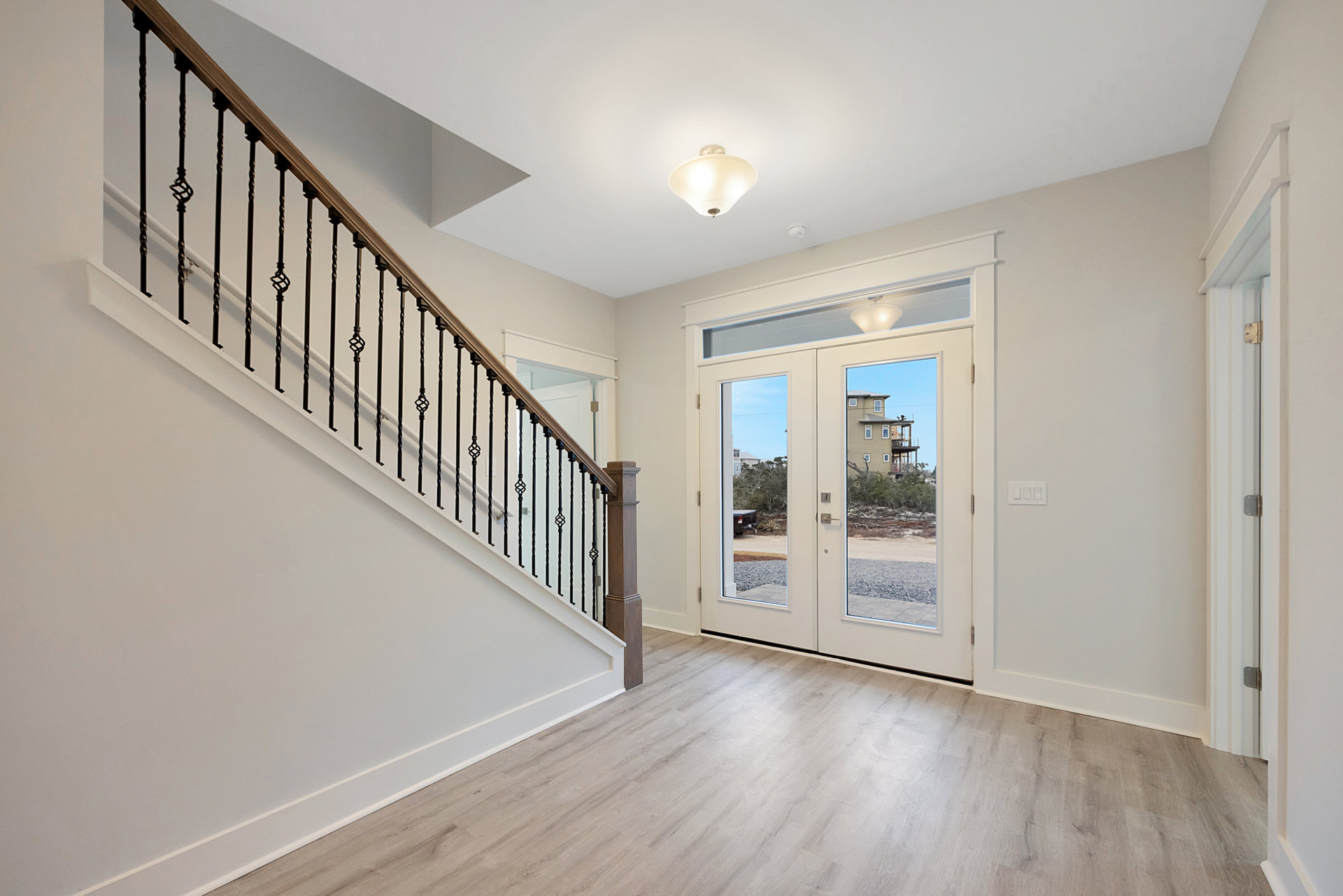 White wood flooring and metal-railed staircase leading to double doors, with balcony, deck, and tree views visible through glass.