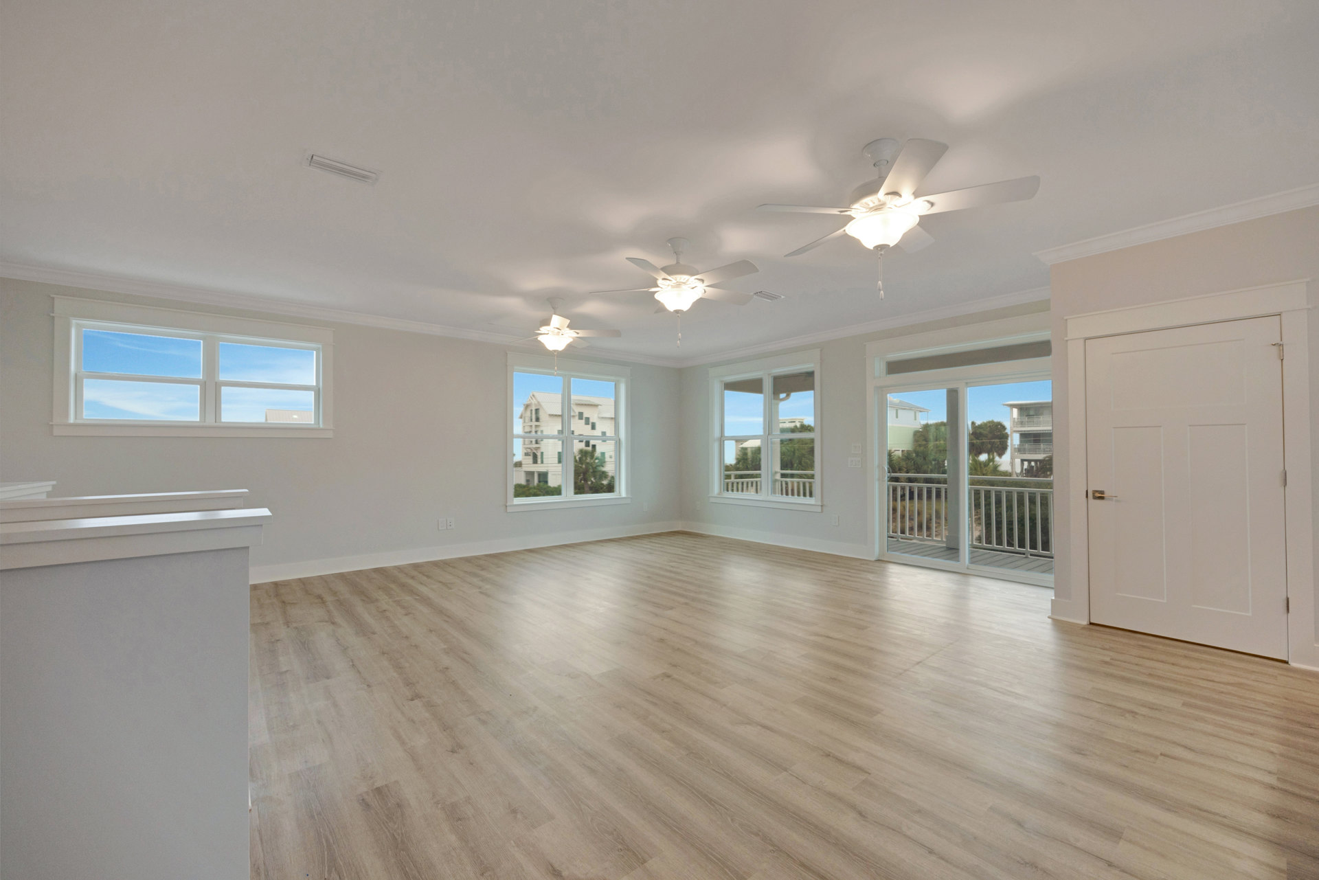 Spacious room with white ceiling fan and light fixture, laminate wood flooring, sliding glass door, two windows showing blue sky and neighboring white building, plaster walls and