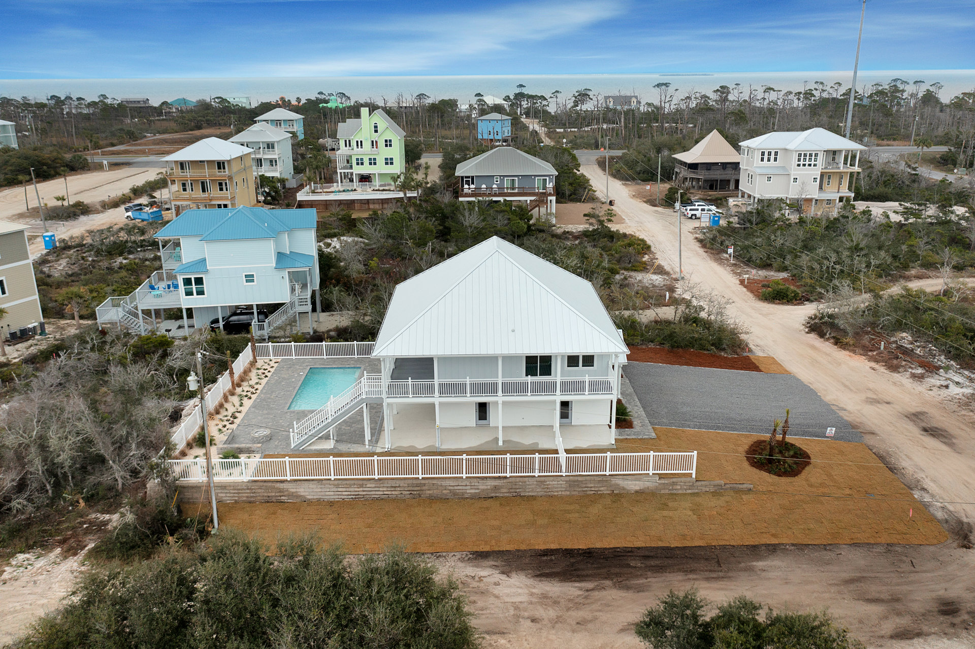 Cluster of modern homes surrounding a central rectangular pool, white and green facades with varied rooflines, balconies, garage with parked car, landscaped trees, blue sky and