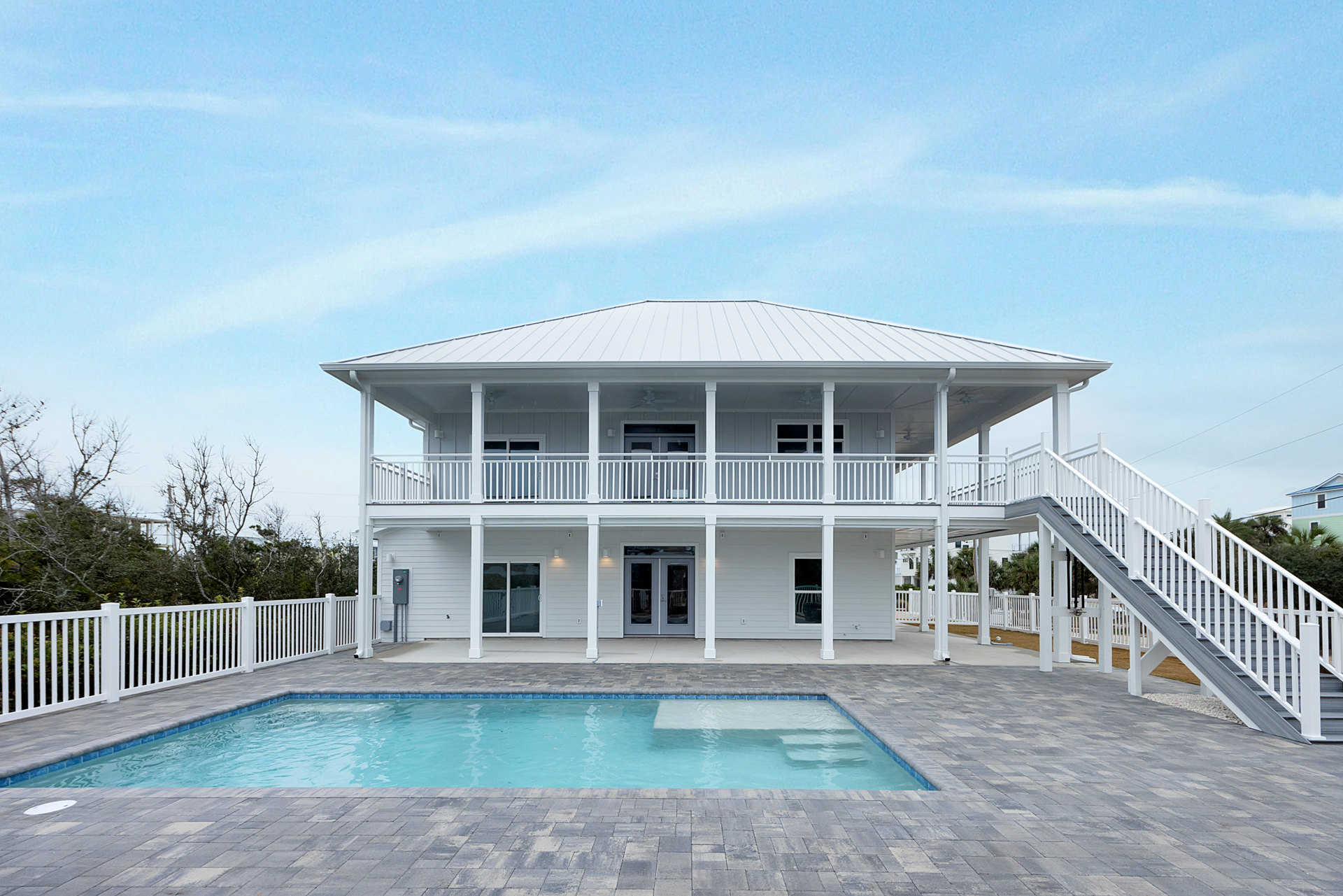 White two-story home with glass balcony doors, tile-edged swimming pool, and white fence surrounded by trees under a clear sky.