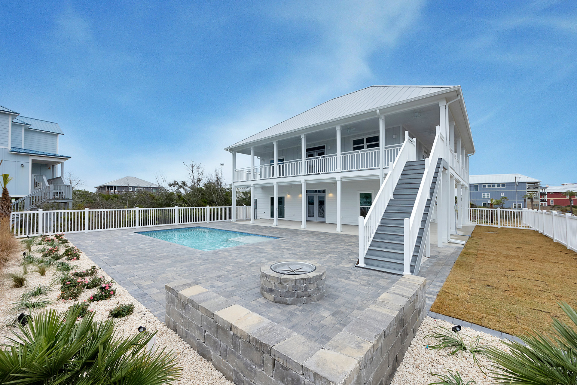 White two-story house with outdoor staircase, rectangular swimming pool bordered by white fencing, circular fire pit on stone patio, manicured lawn, and partly cloudy sky.