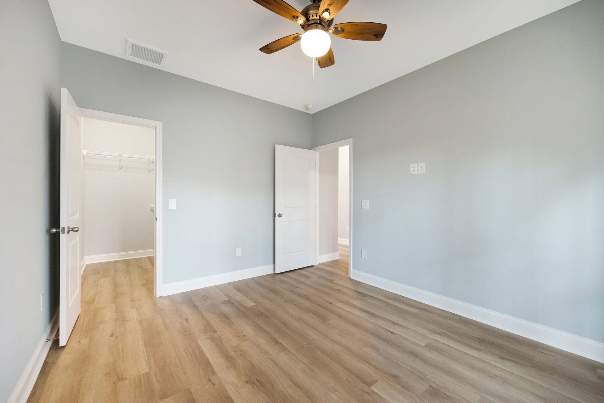 Ceiling fan with light fixture mounted on white plaster ceiling, wood laminate flooring, white door with silver knob, air vent visible near fan.