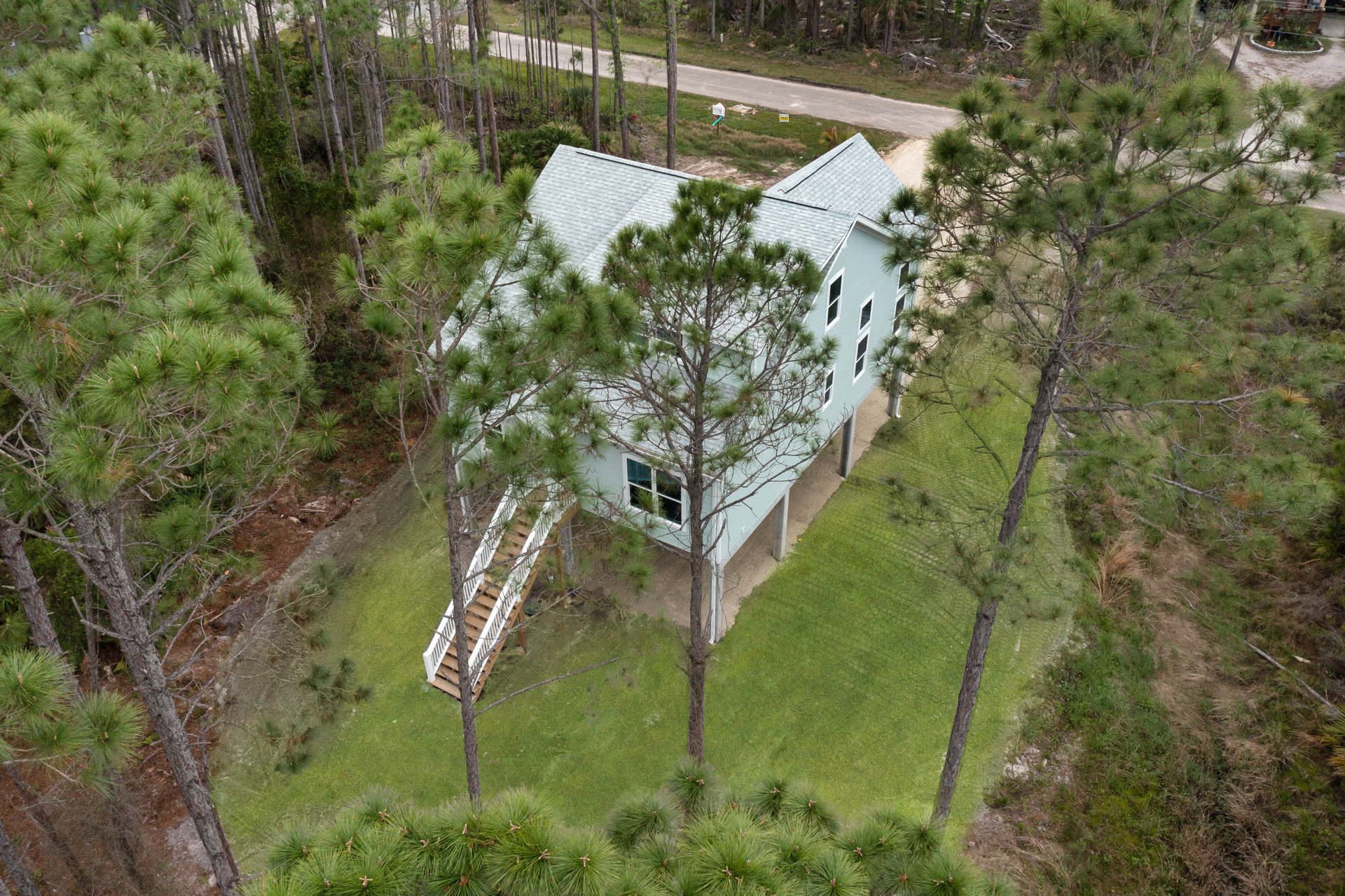 Two-story house with wood siding, white railing staircase, green lawn, and mature trees with leafy and needle foliage on a hillside lot