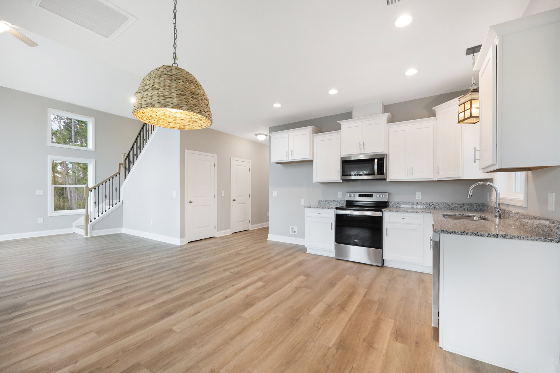 White kitchen cabinets, wood flooring, black and silver oven, built-in microwave, woven pendant light fixture, recessed ceiling light, window with view of trees.