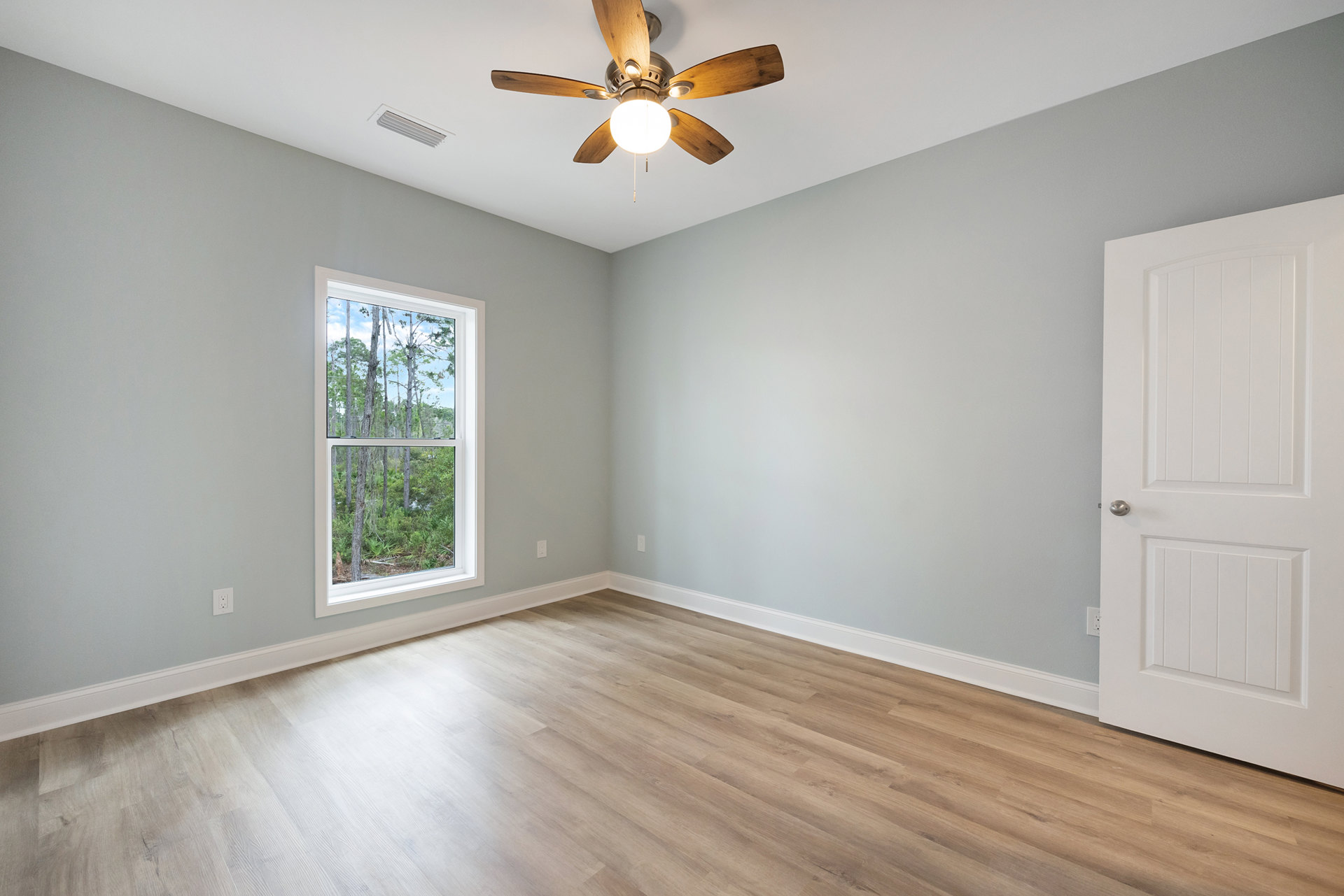 Wood floor room with white walls, ceiling fan with light, large window showing trees outside, and white door with silver handle