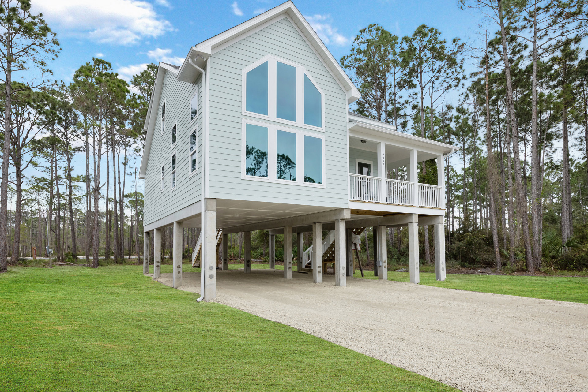 Elevated house on concrete stilts with white porch railing, blue door, gravel path, grassy yard, and trees visible through windows in the background