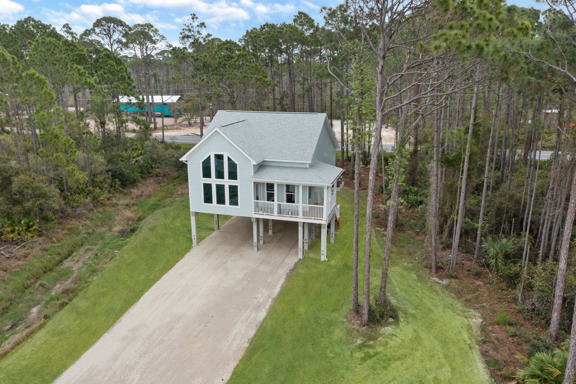 Modern two-story house with light siding and large windows, surrounded by mature trees and landscaped grass, concrete driveway leading to entrance