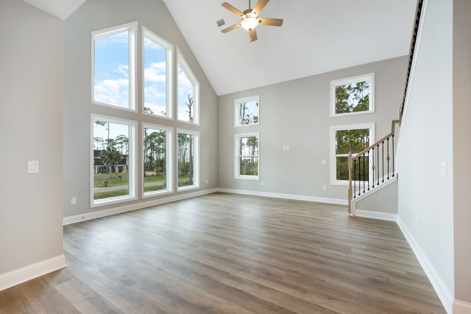 Ceiling fan with light fixture above hardwood floor, large windows with tree views, staircase with wood railing, sunlight streaming across laminate flooring