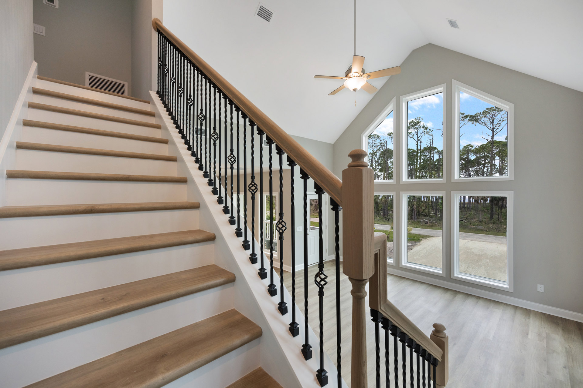Wood staircase with matching railings, balusters, and treads, ceiling fan with light fixture above, neutral walls and flooring, natural light from nearby window.