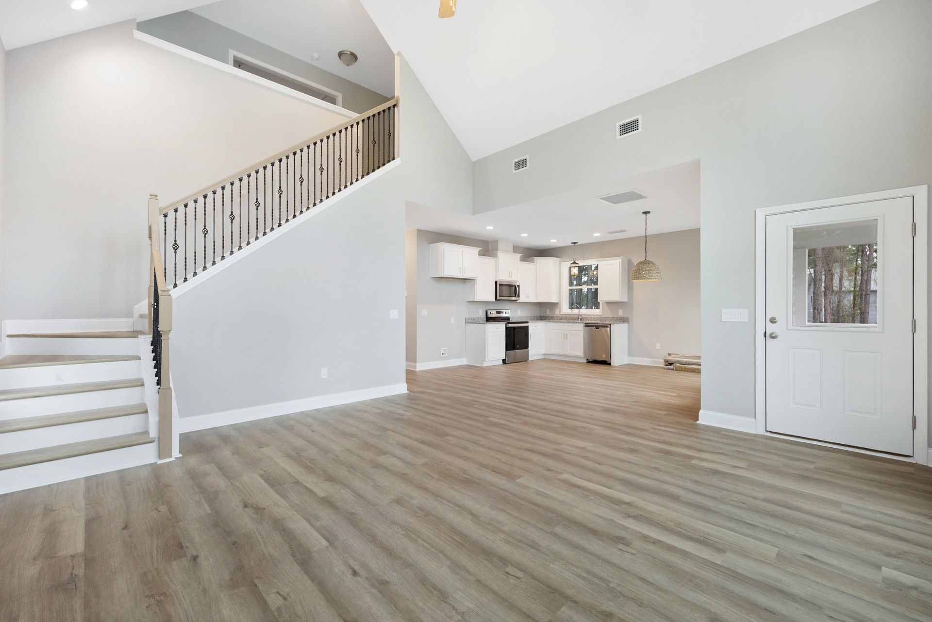 Open-concept room featuring wood flooring, white staircase with wood railing, modern kitchen with stainless steel oven, white cabinetry, and a window overlooking trees.