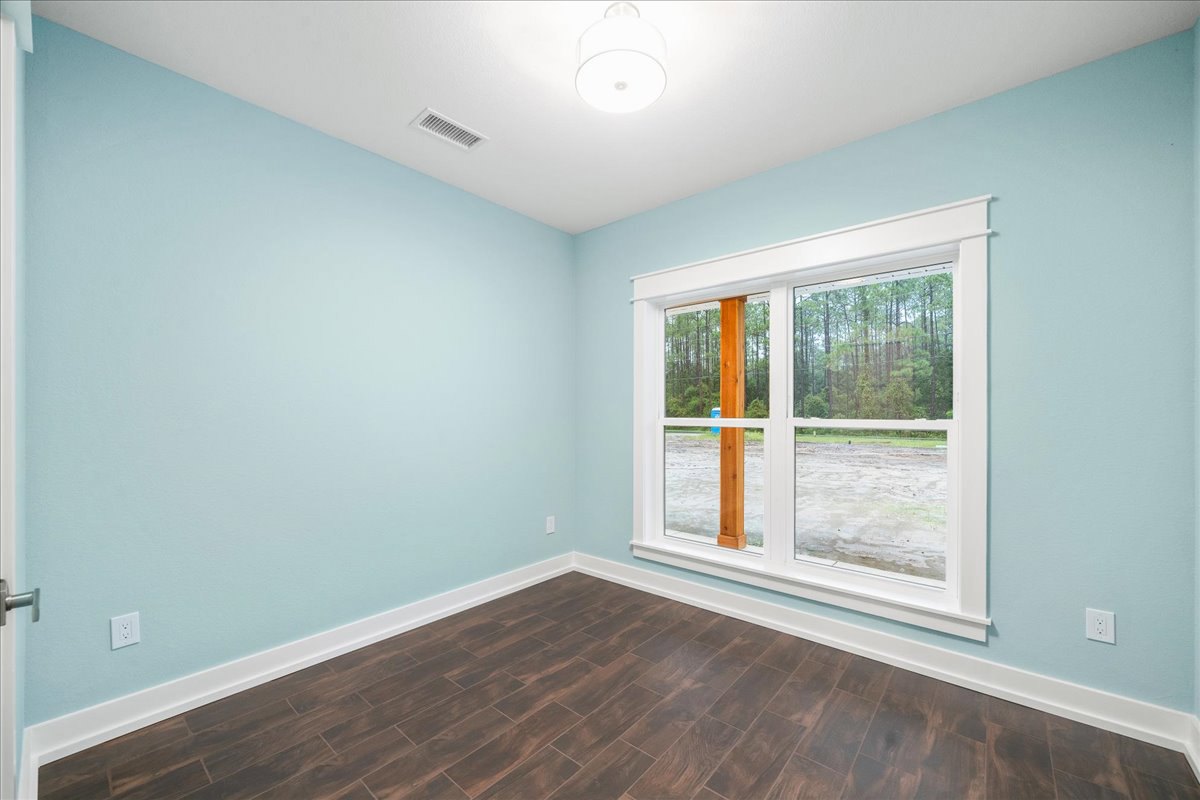Sunlit room featuring wide wood flooring, white baseboards, a large window framed by a wood beam, white ceiling with a modern light fixture, wall vent, and leafy trees visible