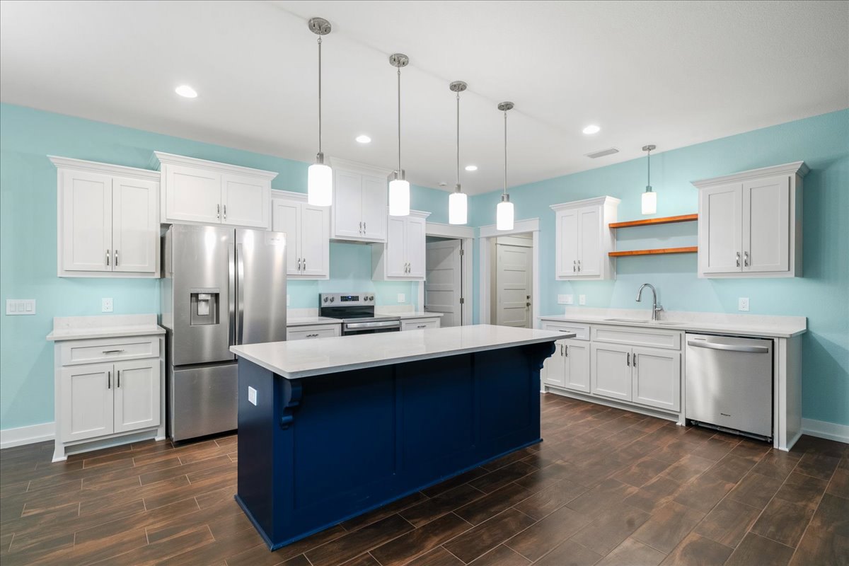 Blue kitchen island with white countertop, stainless steel refrigerator and dishwasher, white cabinetry with silver handles, light wood flooring, and modern fixtures.