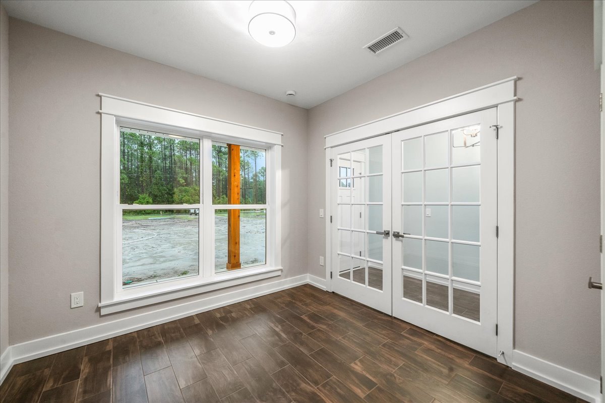 Wood floor with white trim, double glass-paneled doors, large window overlooking dirt road and trees, white ceiling vent, and modern white light fixture with central opening