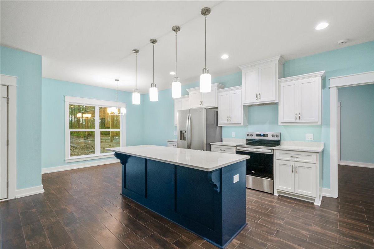 Blue and white kitchen island with quartz countertop, stainless steel refrigerator and stove, white cabinets with silver handles, tile backsplash, large window overlooking outdoor