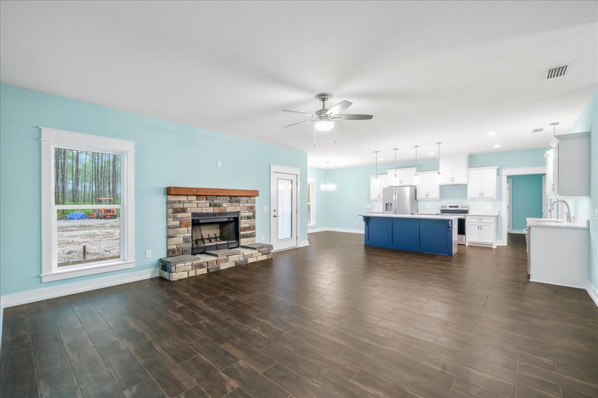Open living room with wood flooring, black-framed fireplace, ceiling fan with light, adjacent kitchen, large window showing tractor outside